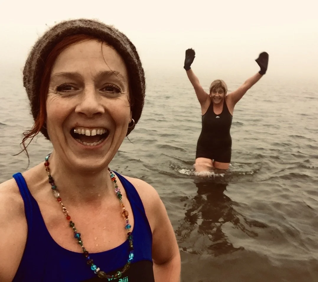 Two women smiling in a body of water, one in the foreground taking a selfie, the other in the background with arms raised, both wearing black swimsuits.