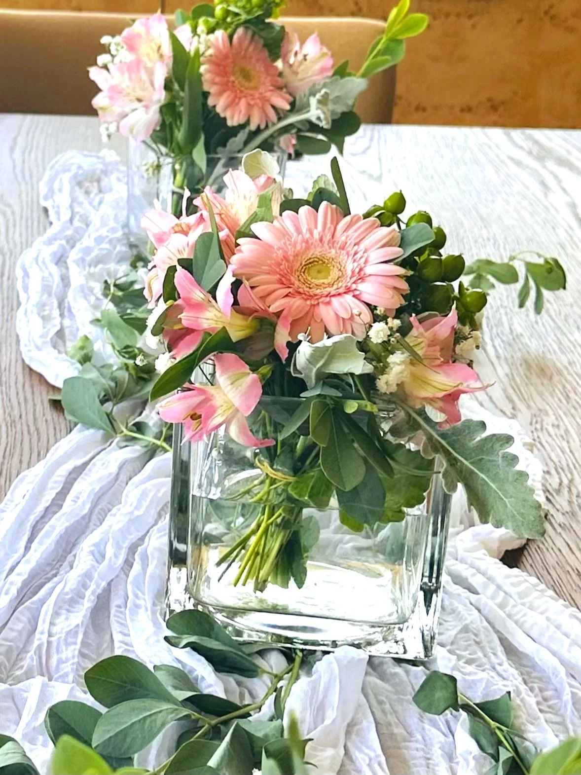 Pink and white flowers in a glass vase on a white cloth on a wooden table.