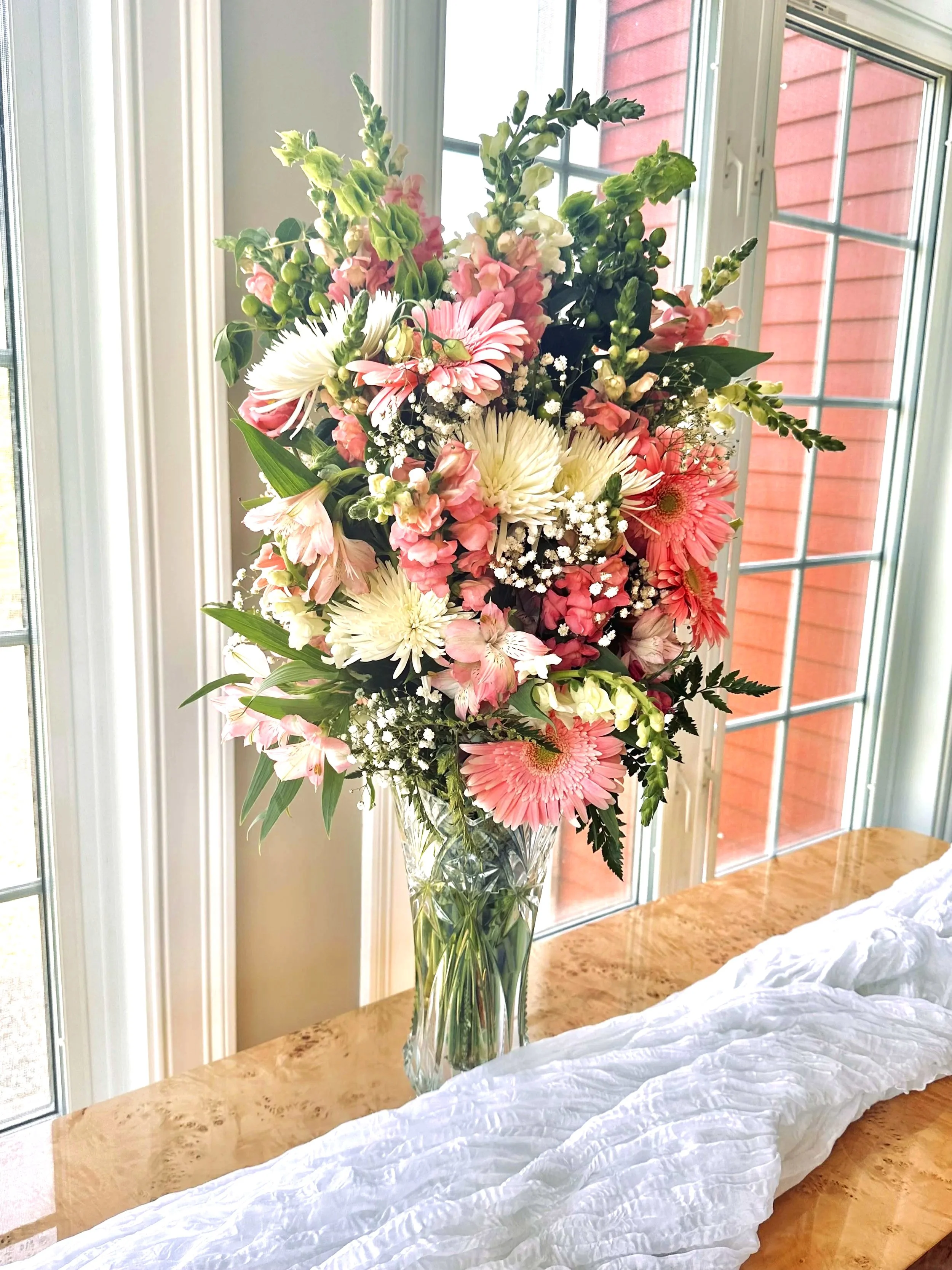 A tall, clear glass vase with a large arrangement of pink and white flowers, including gerbera daisies, chrysanthemums, snapdragons, baby's breath, and greenery, on a wooden table near a window.