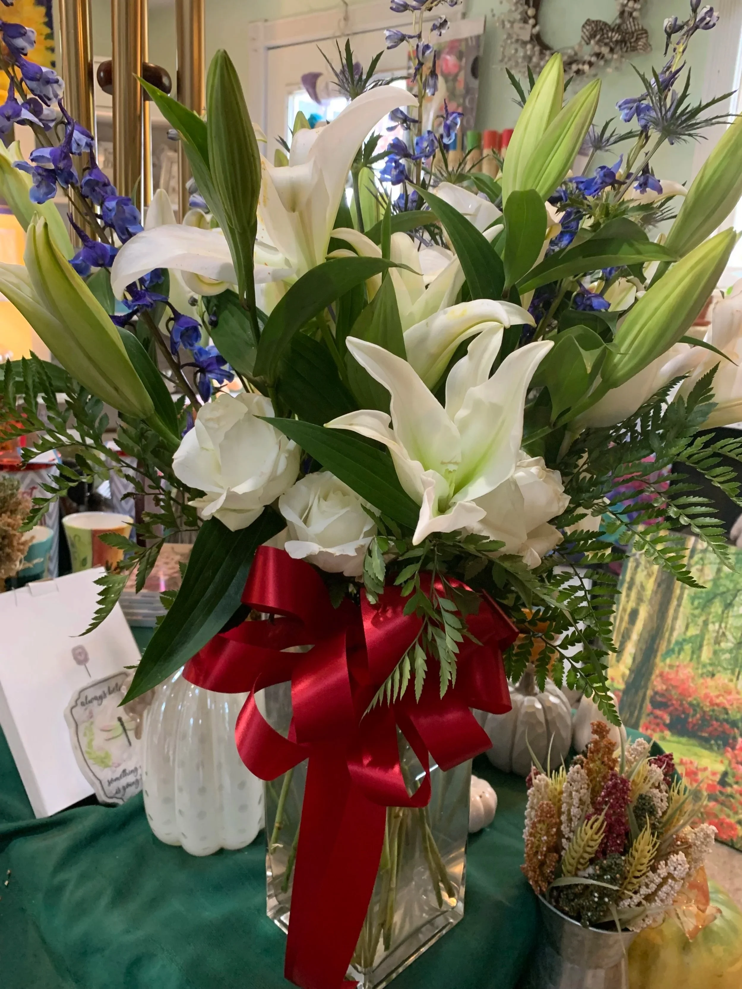 A bouquet of white lilies and white roses, accented with greenery and tied with a large red bow, arranged in a tall clear glass vase.
