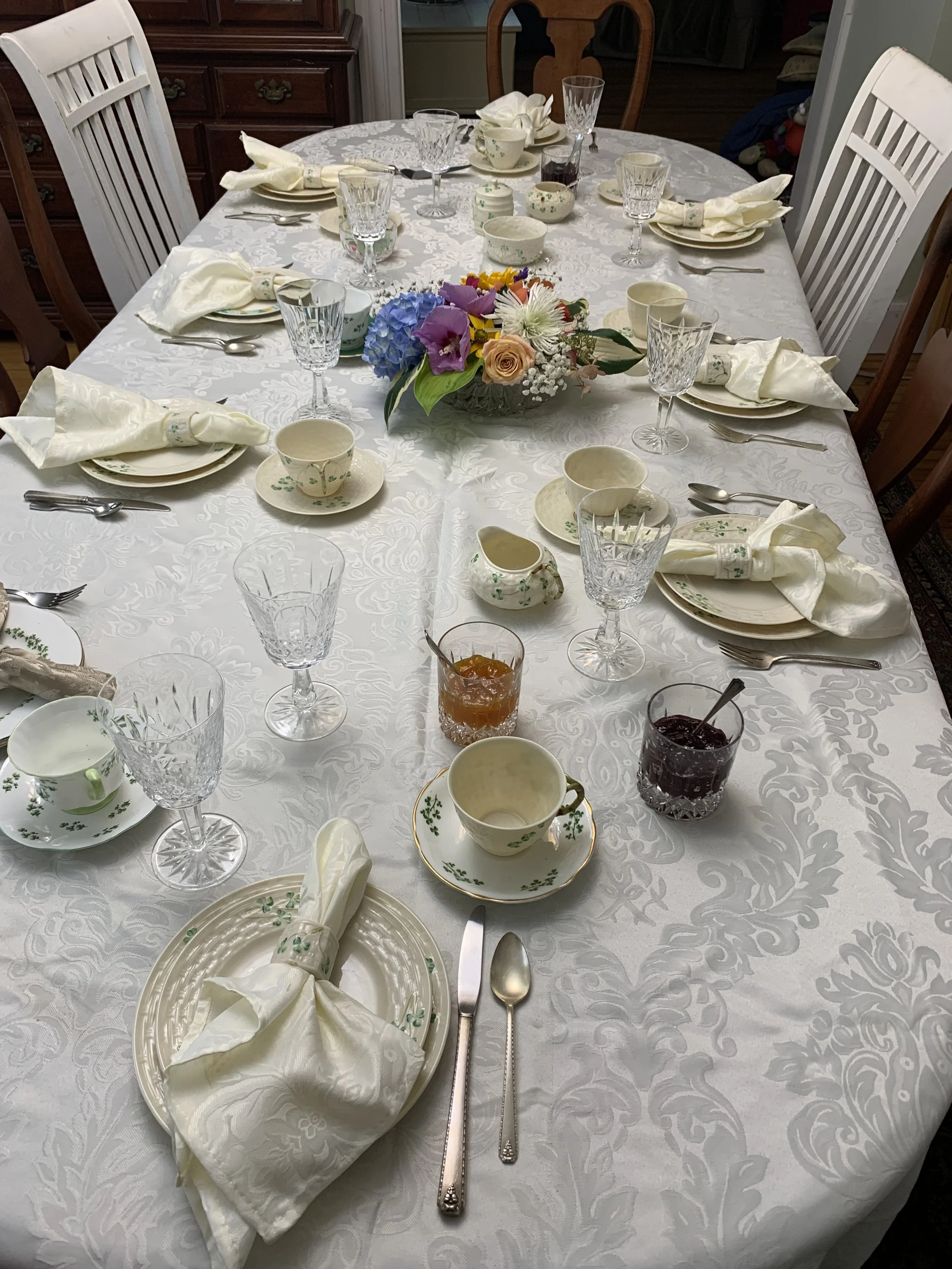 A decorated dining table set for a meal with white floral tablecloth, fine china, crystal glassware, silverware, and a floral centerpiece.