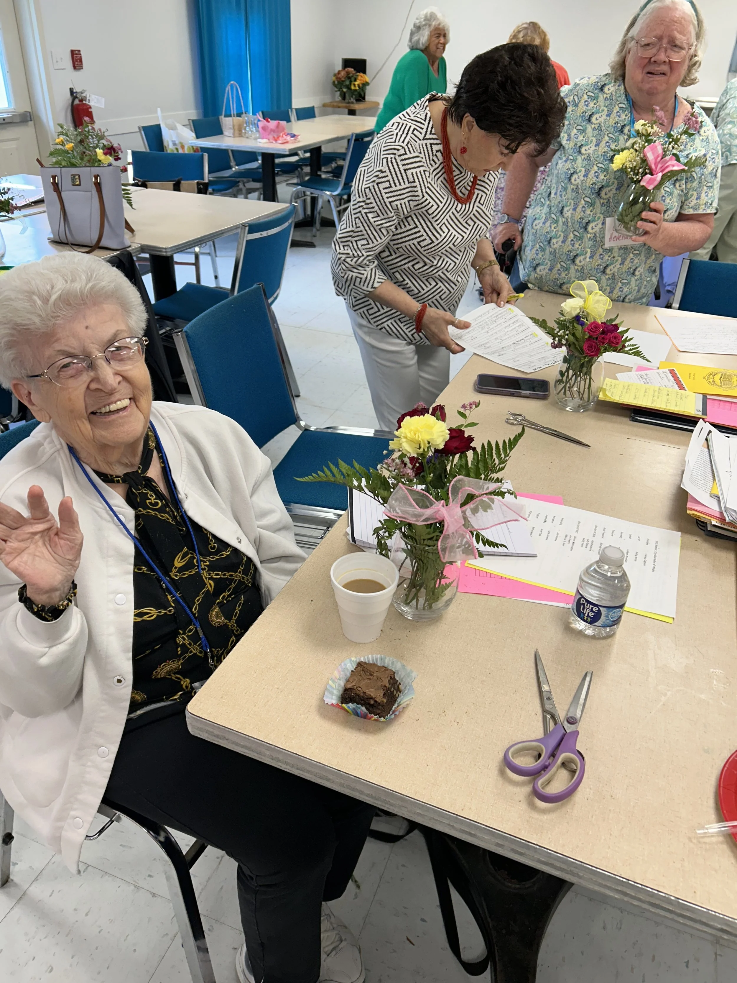 Group of elderly women gathered around a table decorated with flowers, papers, and snacks celebrating an event or special occasion.