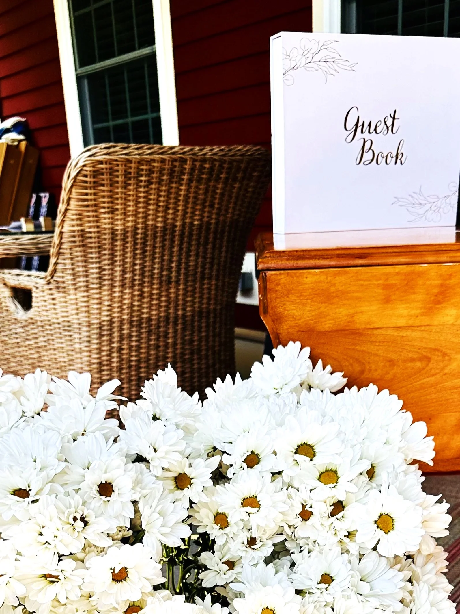 A white guest book on a wooden table next to a bouquet of white daisies with yellow centers, on a porch with a wicker chair and a red exterior wall in the background.