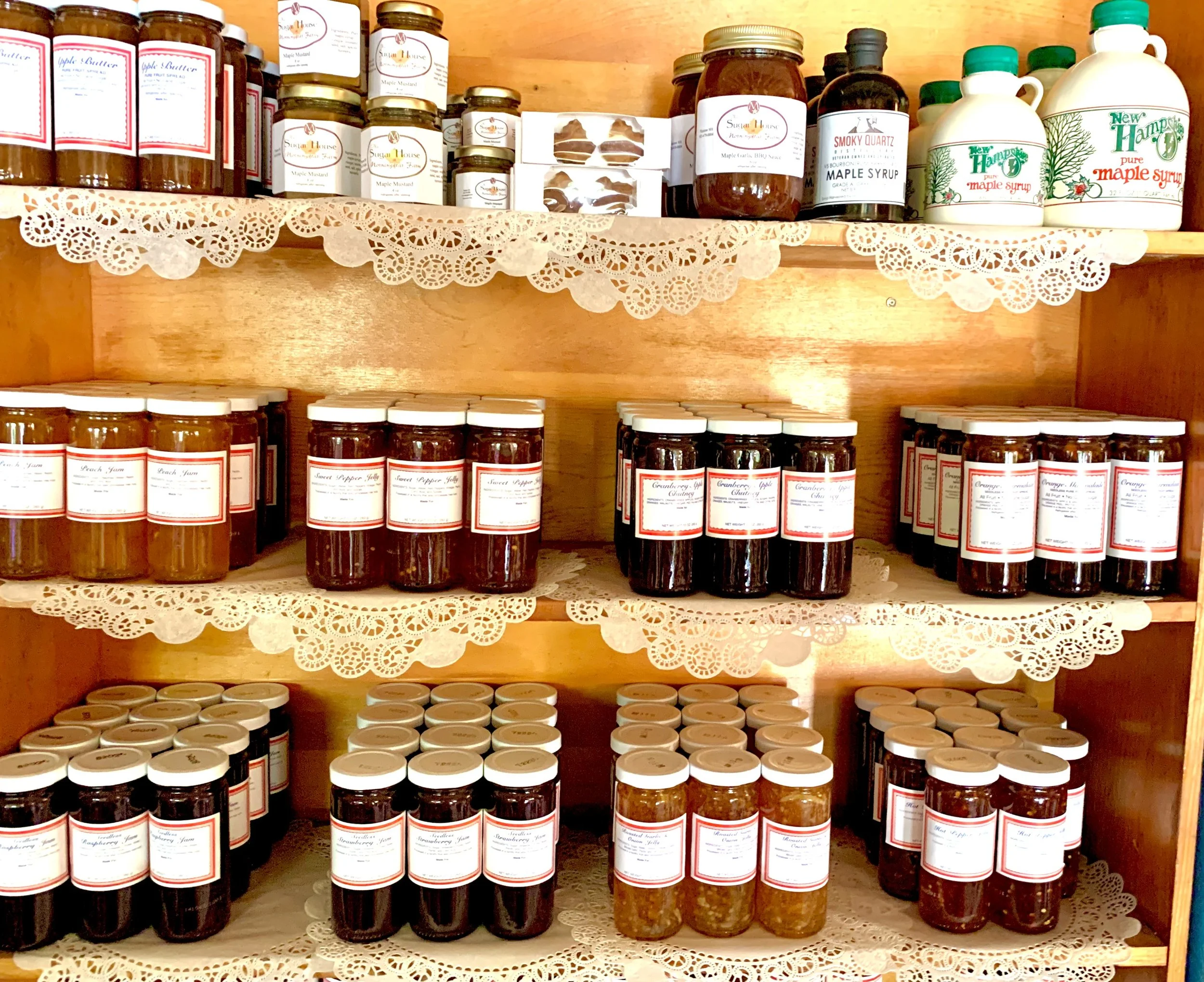 Wooden shelf filled with jars of homemade preserves, jams, and syrups, arranged on lace doilies, with bottle labels indicating flavors and ingredients.