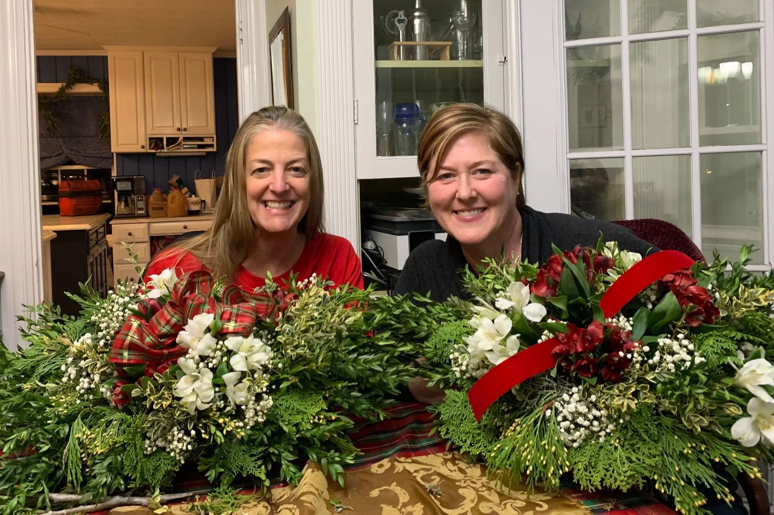 Two women smiling behind two large holiday floral arrangements on a table inside a house.