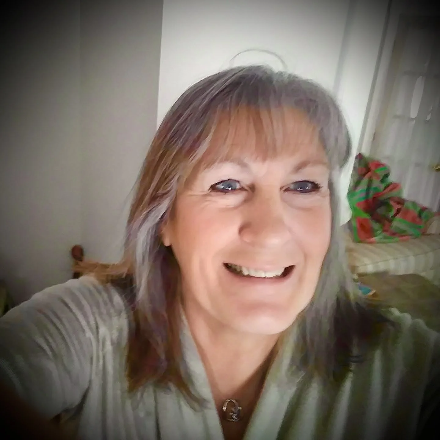Close-up of a smiling middle-aged woman with shoulder-length gray hair with purple highlights, blue eyes, and fair skin, taken indoors with a background of a sofa with Christmas pillows and a white door.