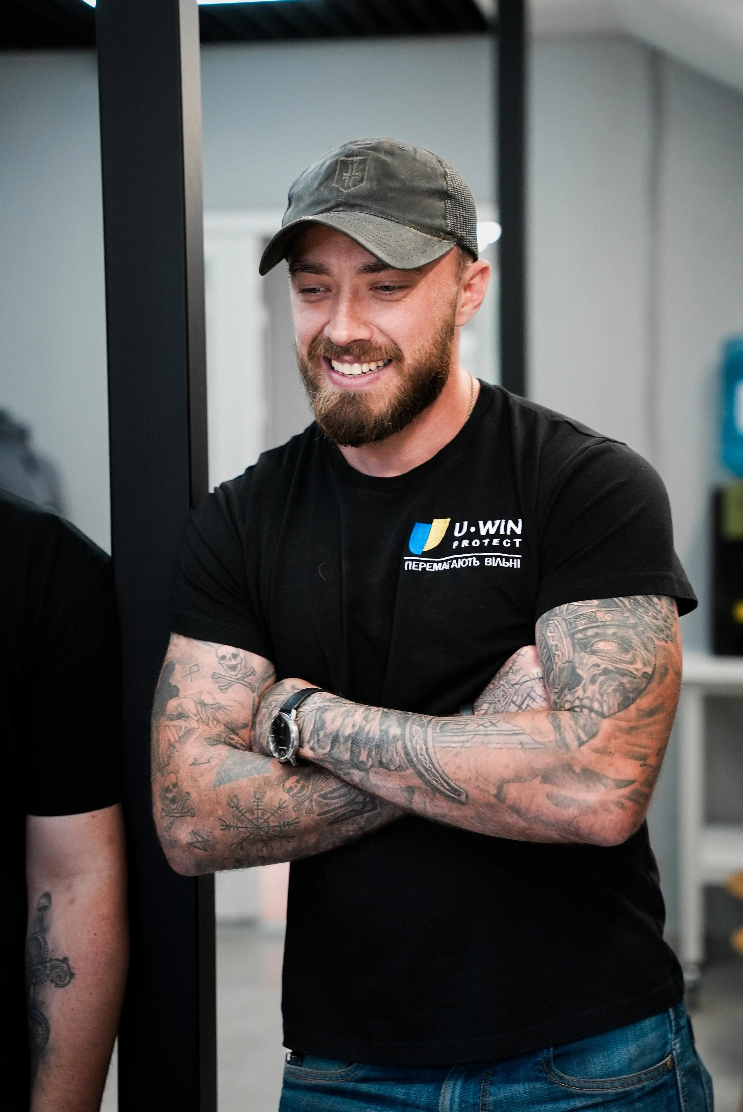 Young man with a beard, tattoos on arms, wearing a black T-shirt with a logo, gray cap, and watch, smiling with arms crossed.