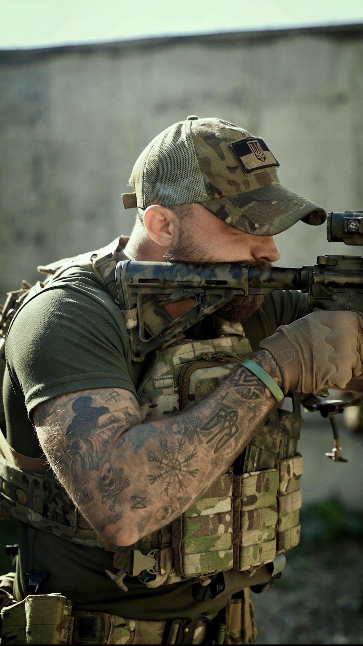 A soldier in camouflage uniform aiming through a scope with a rifle, wearing a camo cap and tactical vest, with tattoos on his arm.