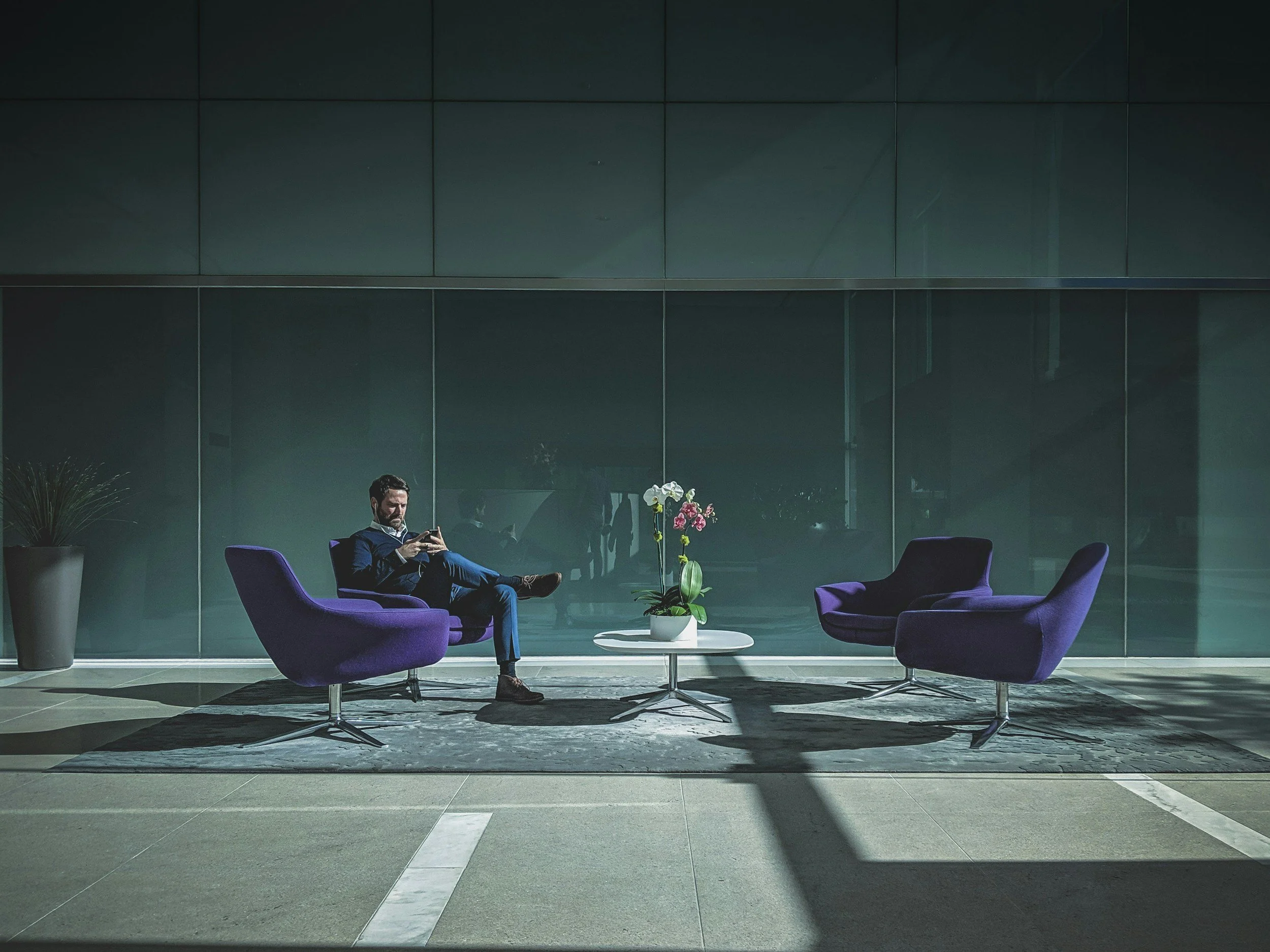 A man in a suit sitting on a purple chair in a modern, minimalistic lobby, looking at his phone with two additional purple chairs, a white table with orchids, and a large potted plant in the background.