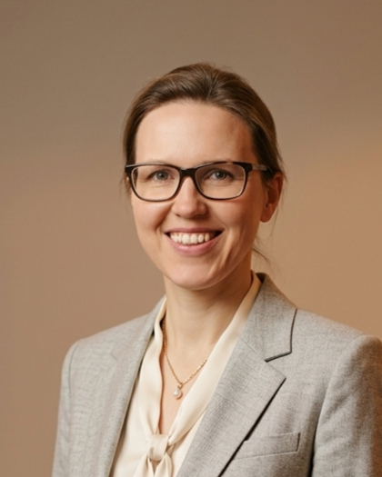 Porträt einer lachenden Frau mit Brille in formeller Kleidung vor einem neutralen Hintergrund.
