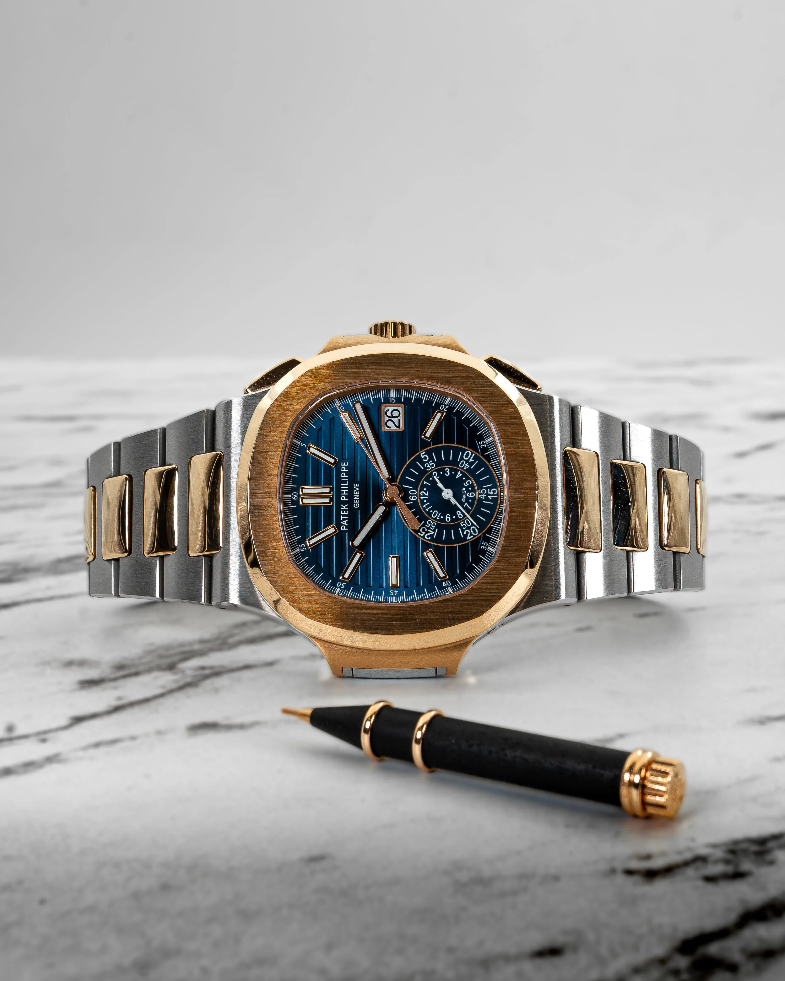 A luxury wristwatch with a blue dial, gold bezel, and two-tone metal band, positioned on a marble surface alongside a black and gold pen.