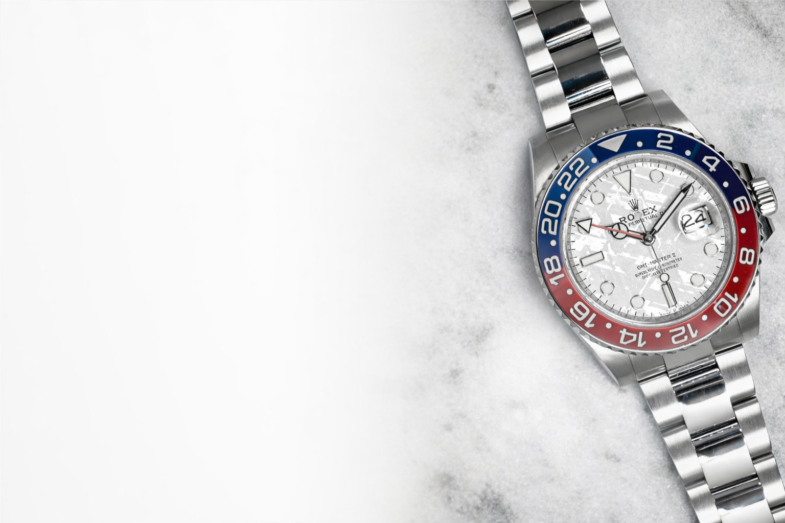 Close-up of a Rolex GMT-Master II watch with a silver band, white textured dial, and red and blue bezel, resting on a gray surface.