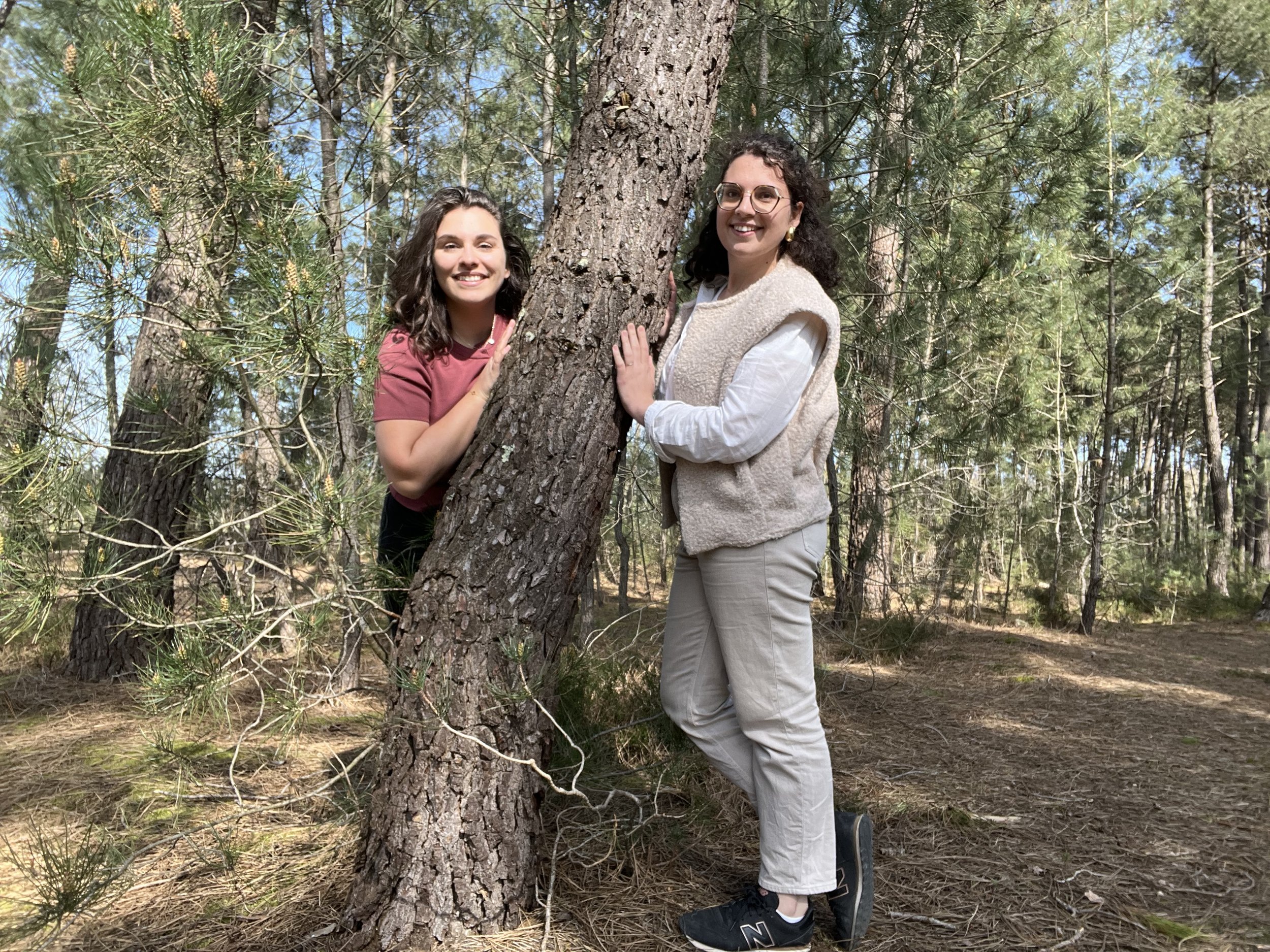 Deux femmes souriantes dans une forêt, l'une tenant un arbre et l'autre le touchant, arborant des vêtements décontractés et des lunettes.