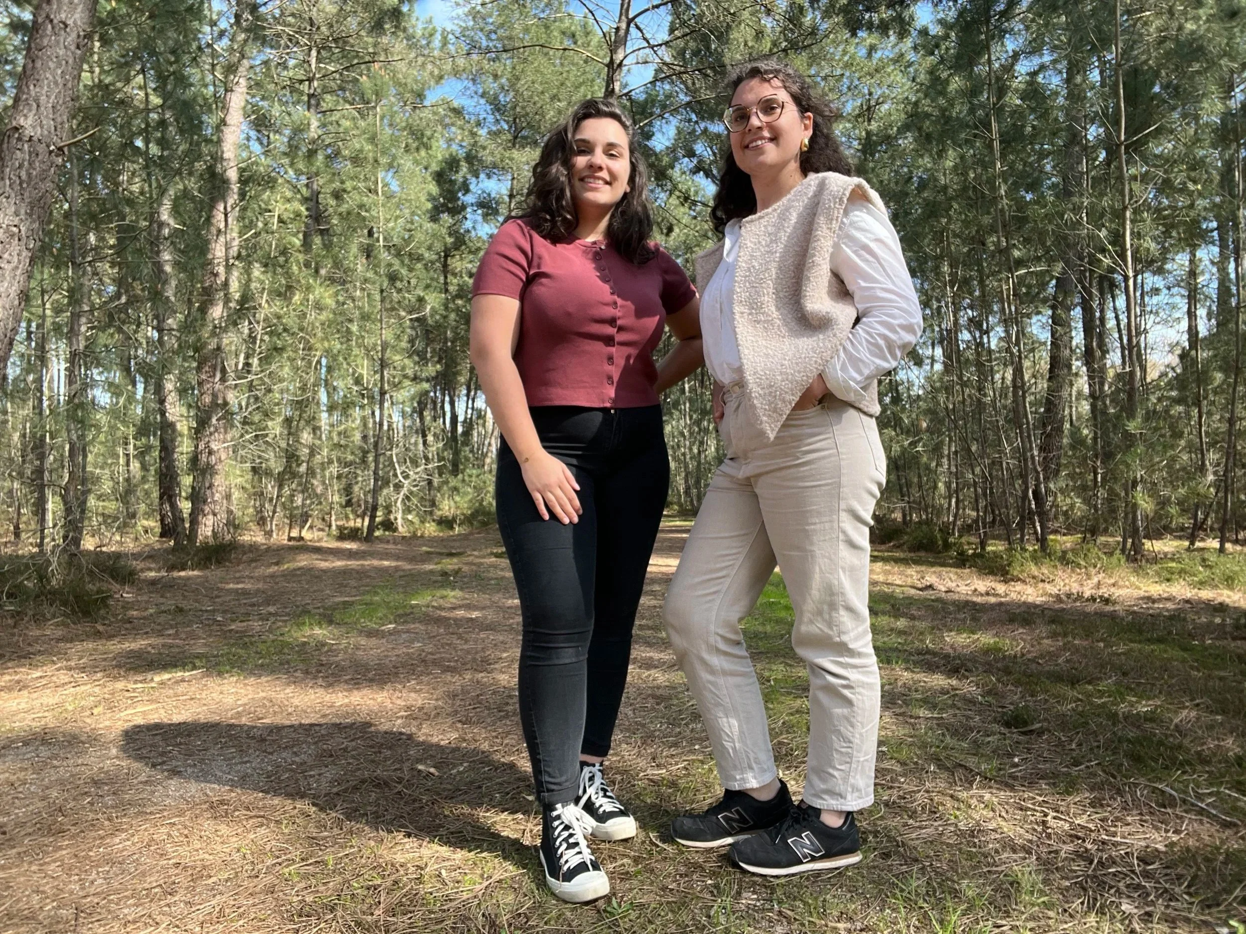 Deux femmes debout dans une forêt ensoleillée, souriantes, portant des vêtements décontractés et des baskets.
