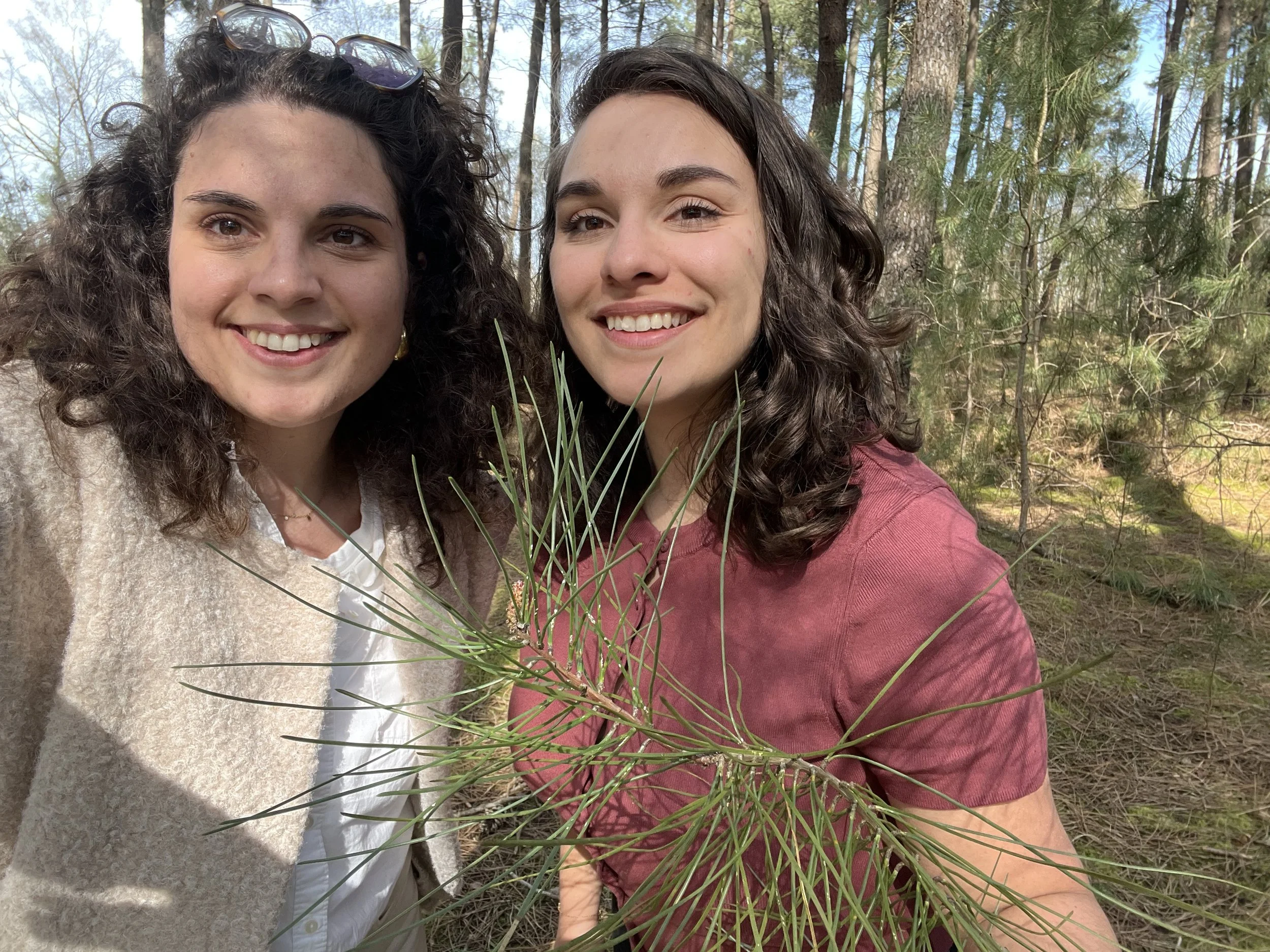 Deux femmes souriantes prennent un selfie dans une forêt, l'une porte une veste beige et l'autre un t-shirt rouge. Elles tiennent une branche d'arbre verte.