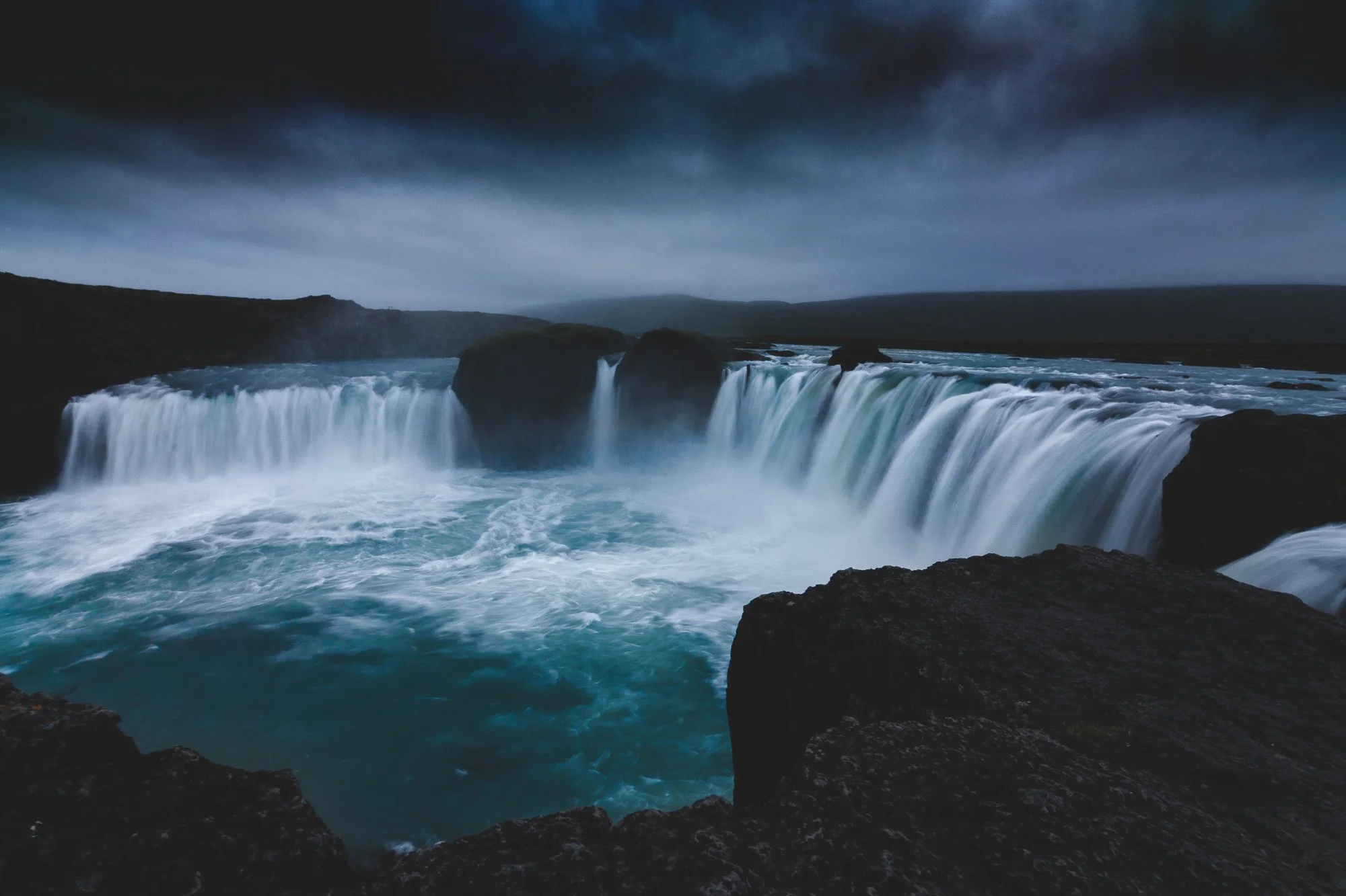 A large waterfall flowing into a turquoise pool under a dark sky, surrounded by rocky cliffs.