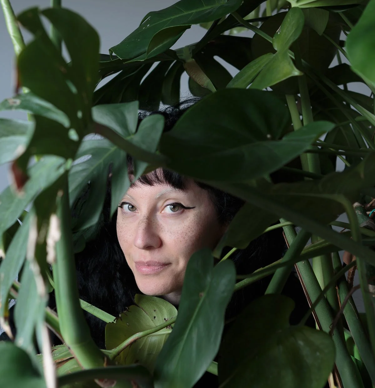 A woman with black hair and freckles peeking through large green plant leaves.