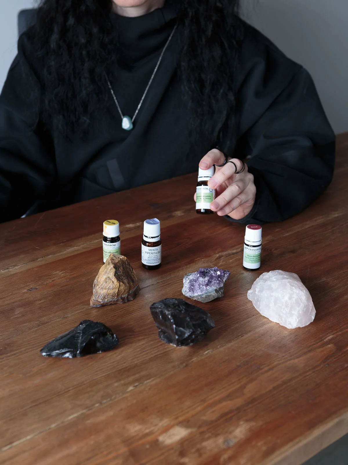 Person with dark curly hair wearing a black shirt and silver necklace sitting at a wooden table with various crystals and essential oils.