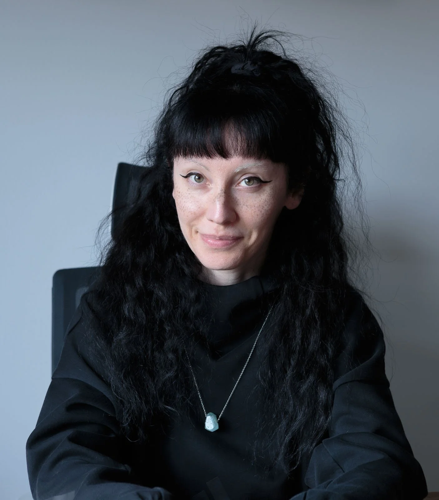 A woman with long, curly black hair, wearing a black top and a necklace, sitting in front of a gray background.