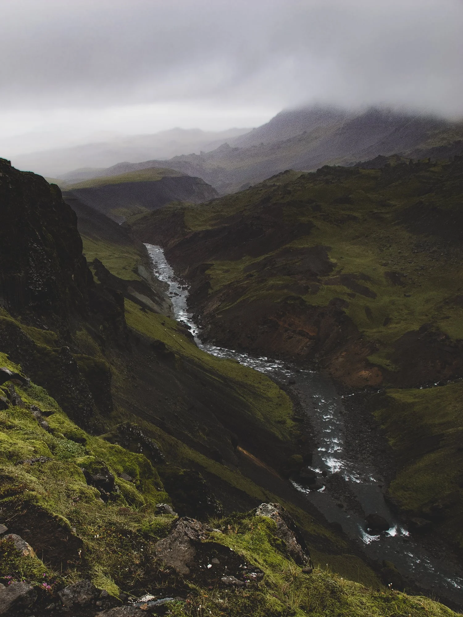 A green and rocky canyon with a river flowing through it, surrounded by hills and mountains under a cloudy sky.
