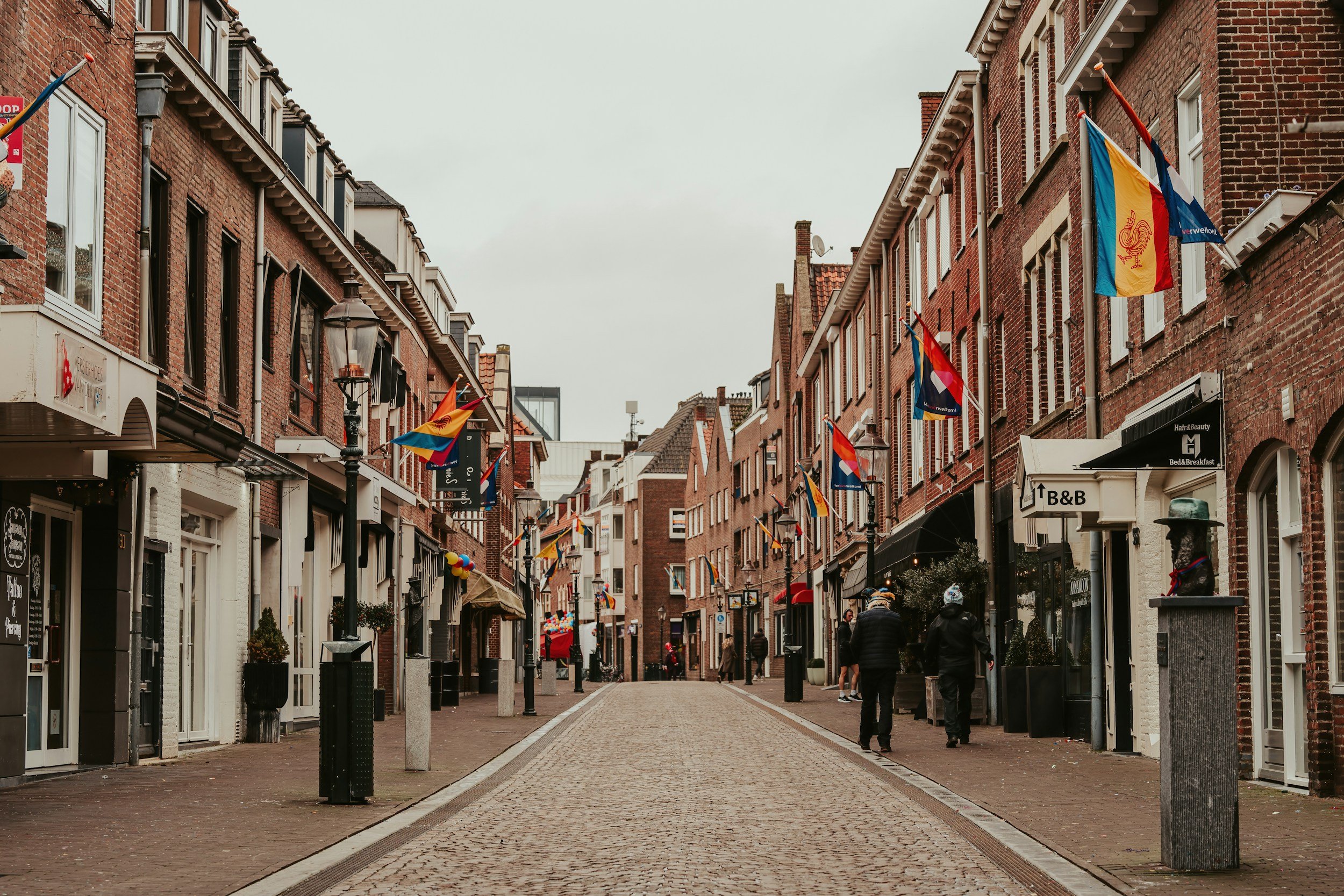 A pedestrian street lined with brick buildings, flags, and shops, with a cloudy sky overhead.
