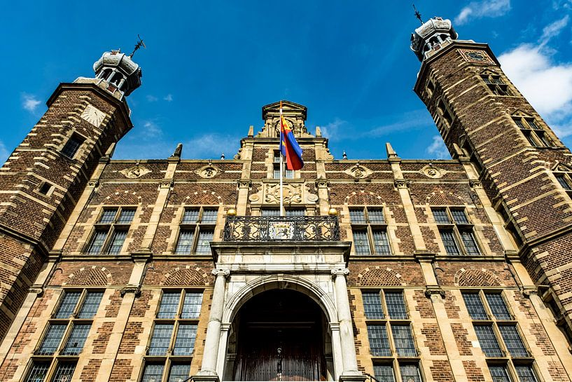 Looking up at a historic castle-like building with two towers, arched entrance, and a flag flying above.