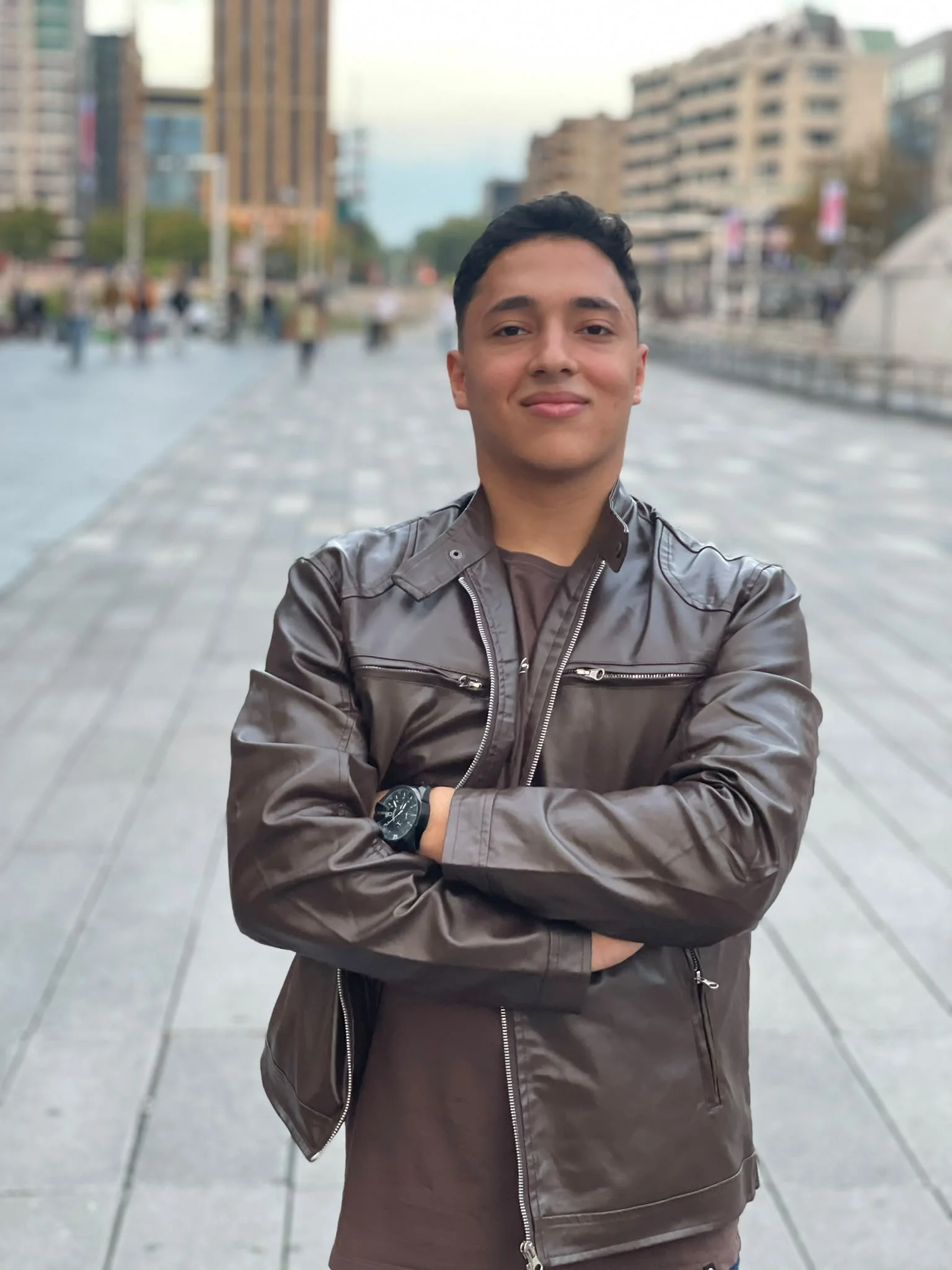 A young man standing outdoors on a city street in daytime, crossing his arms and smiling, wearing a dark brown leather jacket and a watch, with blurred buildings and pedestrians in the background.