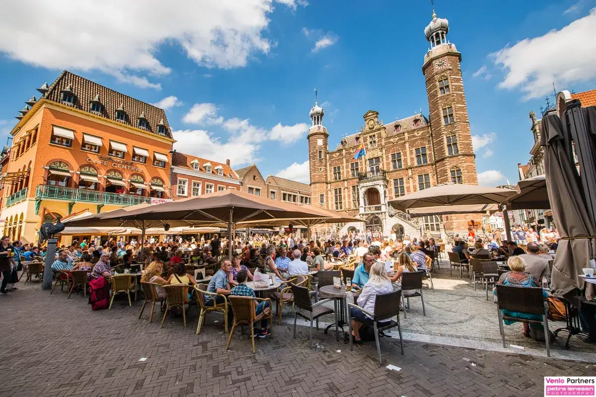 A lively outdoor café scene in a European city square with a historic castle in the background, people sitting at tables under umbrellas, and a clear blue sky.
