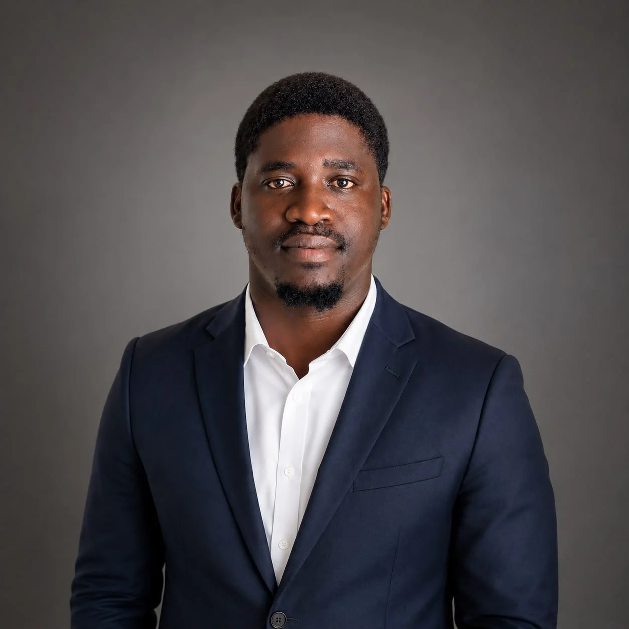 Portrait of a young African American man in a dark business suit and white shirt against a gray background.