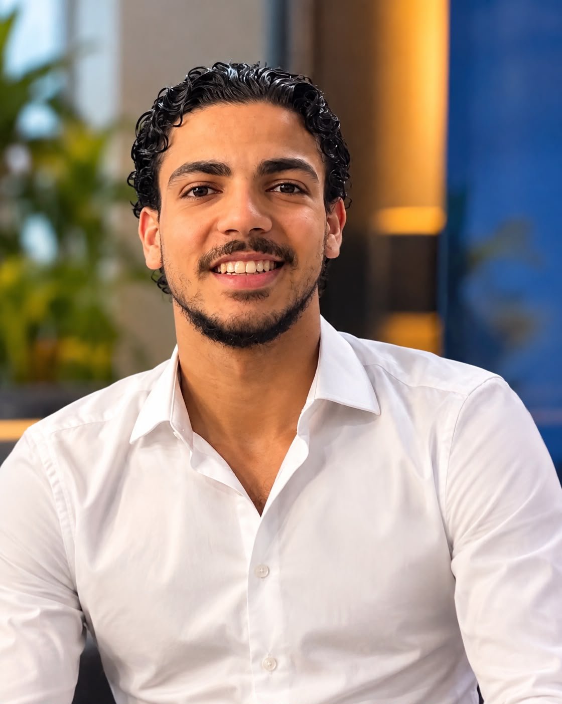 A smiling man with dark curly hair and a beard, wearing a white dress shirt, sitting indoors with a blurred background that includes green plants and a blue wall.