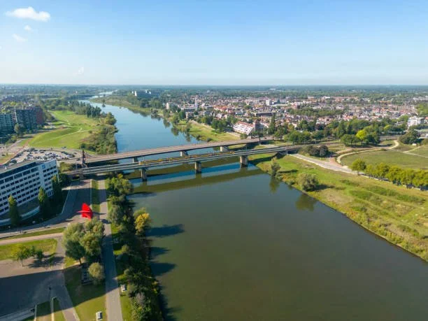 Aerial view of a river with a bridge crossing over it, surrounded by green areas, buildings, and a cityscape in the background under a mostly clear blue sky.