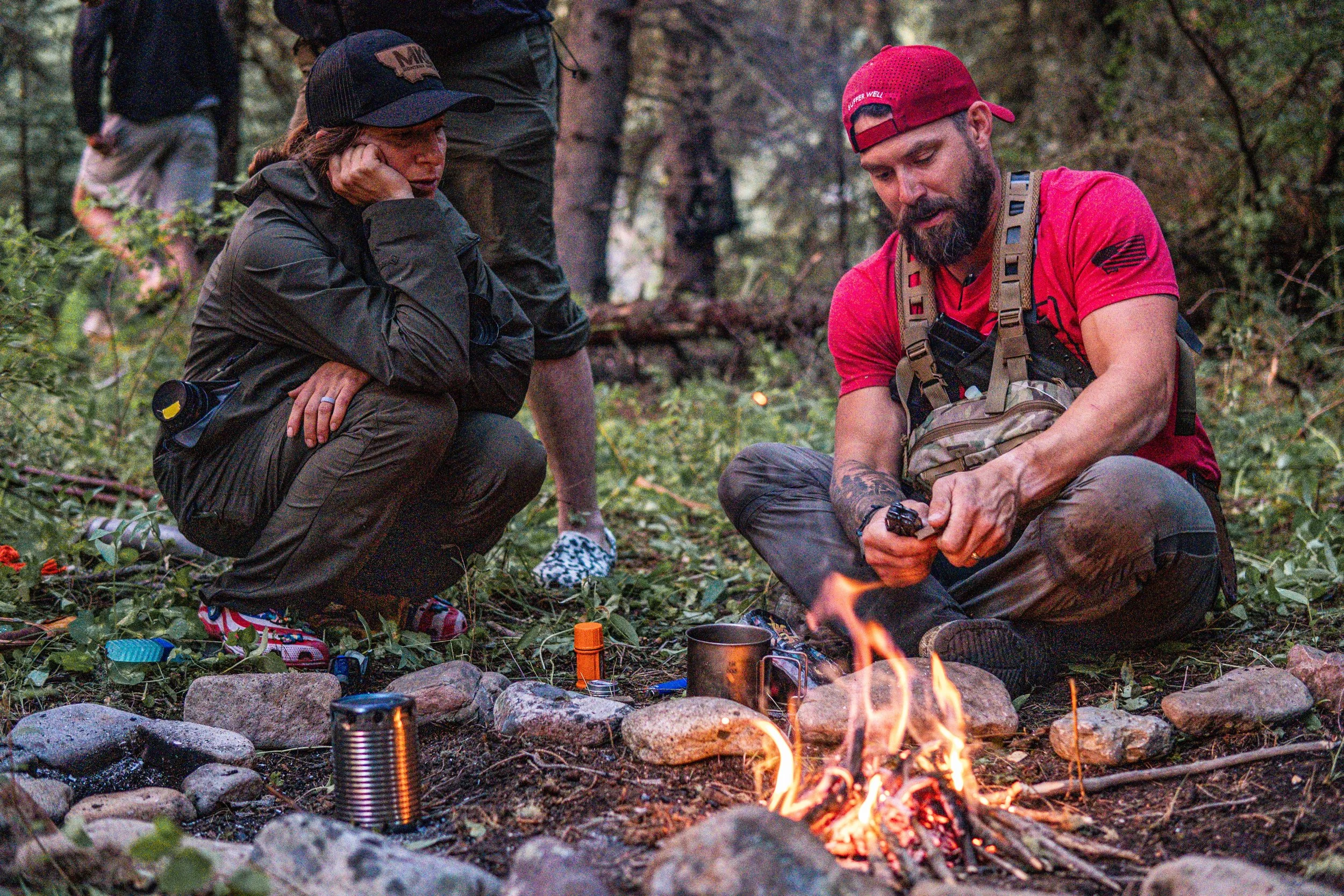 Two people sitting by a campfire in a forested area. One person is wearing a black jacket and cap, resting their chin on their hand, while the other is wearing a red cap and red shirt, tending to the fire. There are trees and foliage in the background.