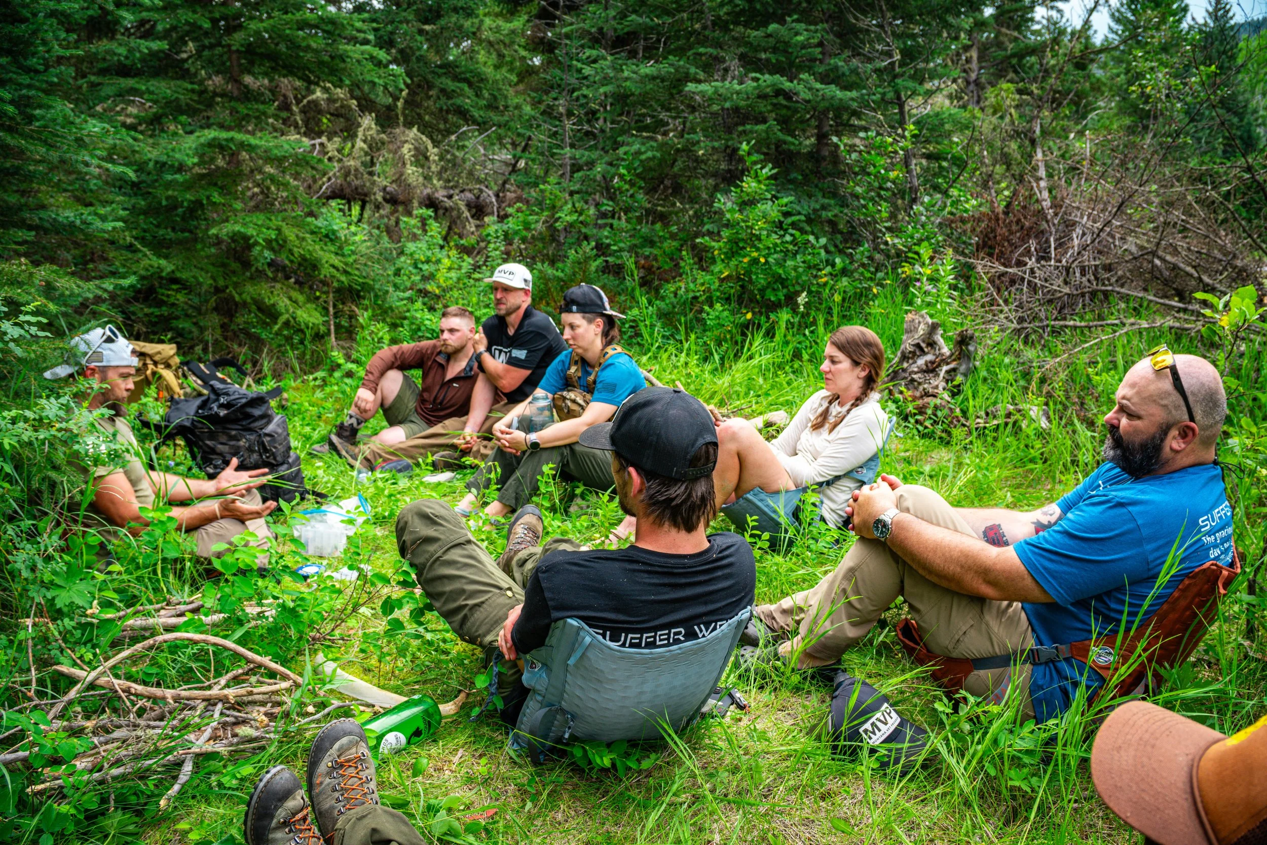 Group of people sitting on the grass in a forested area, engaged in a discussion or meeting, with backpacks and gear around them.
