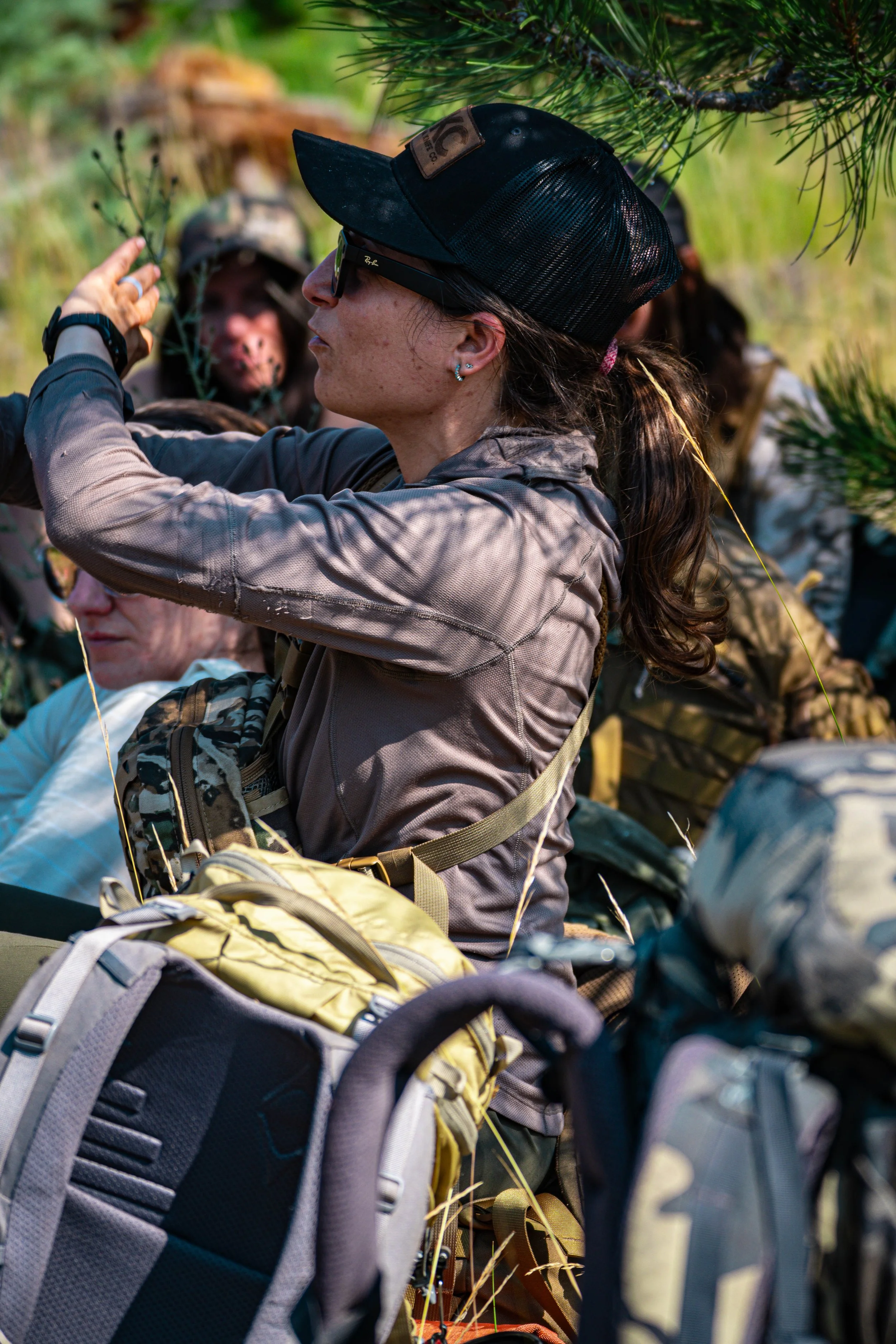 A woman in sunglasses and a baseball cap outdoors with a group, surrounded by backpacks and foliage.