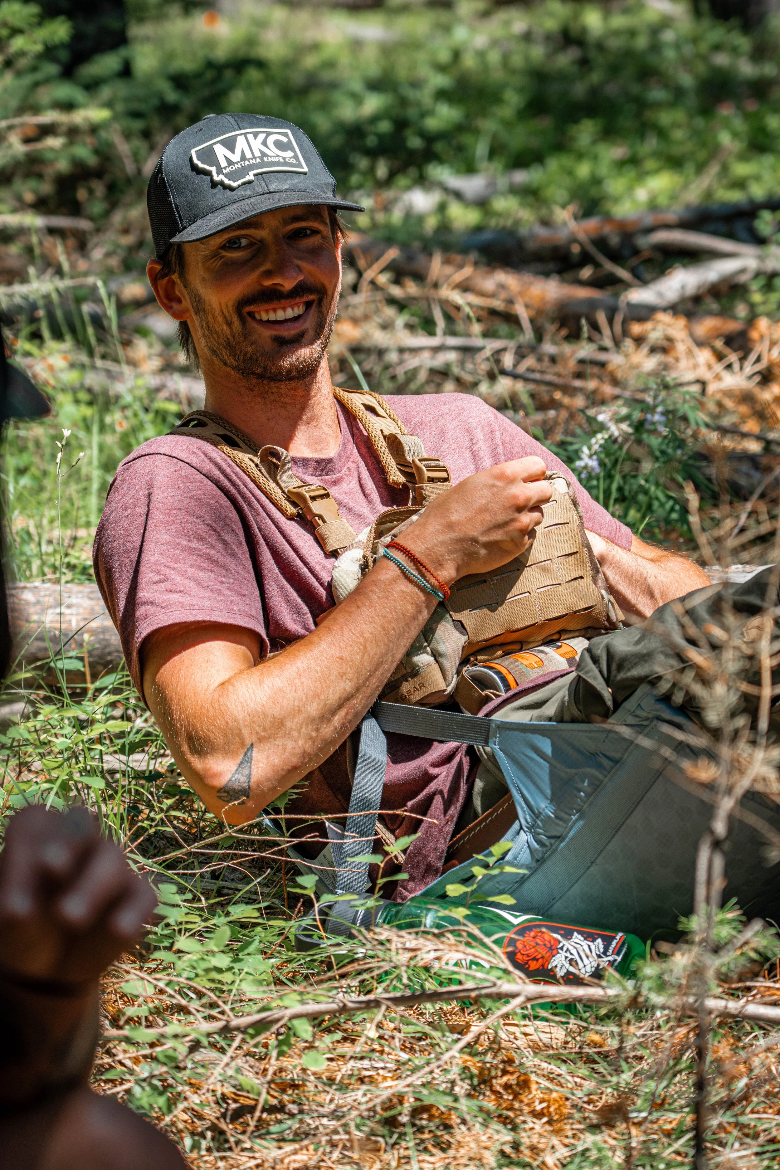 A man smiling, sitting outdoors in a wooded area, wearing a black cap and a camouflage tactical vest.