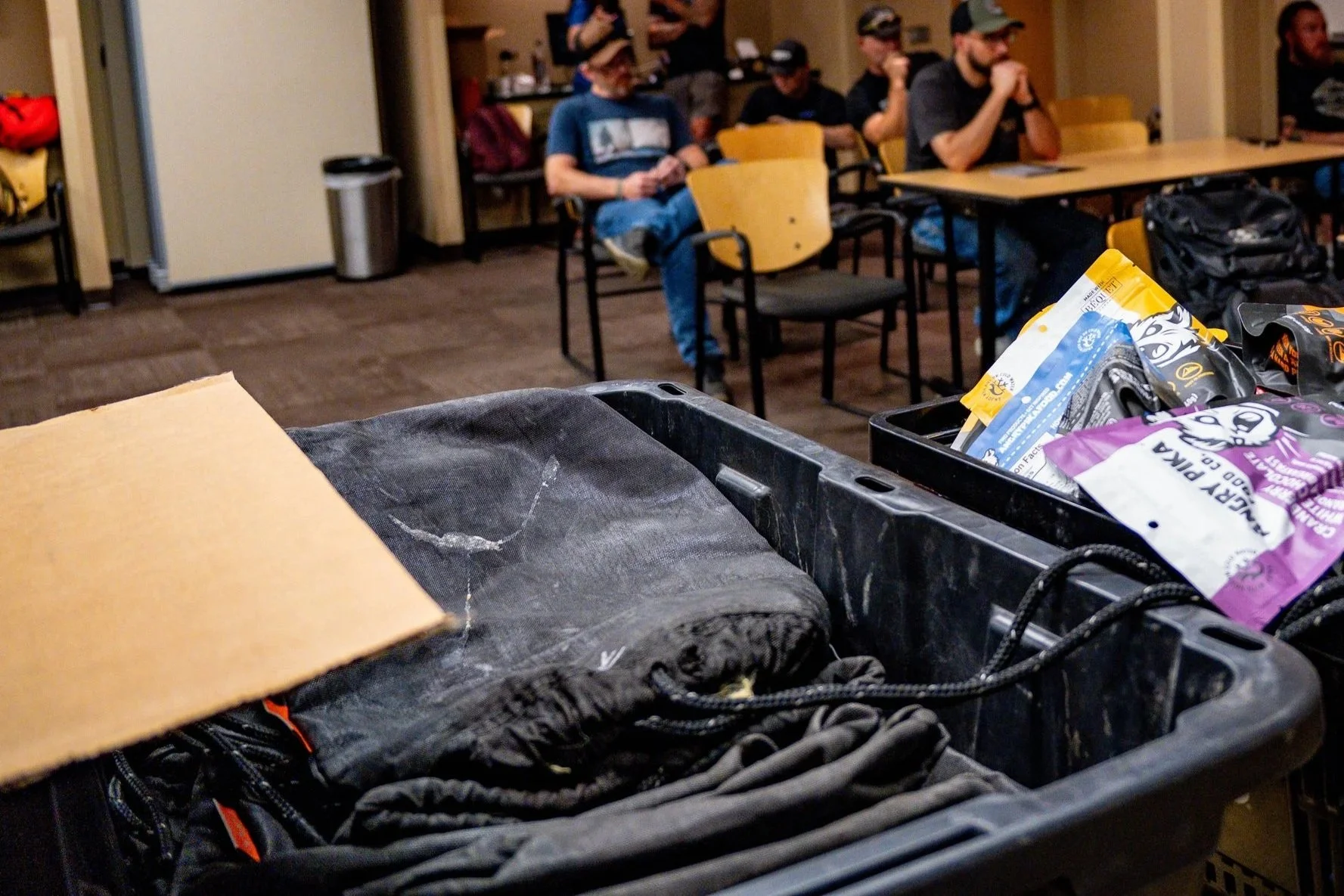 A table with various black bags and snacks in the foreground, and people sitting at tables in a room with wood flooring and a beige wall in the background.