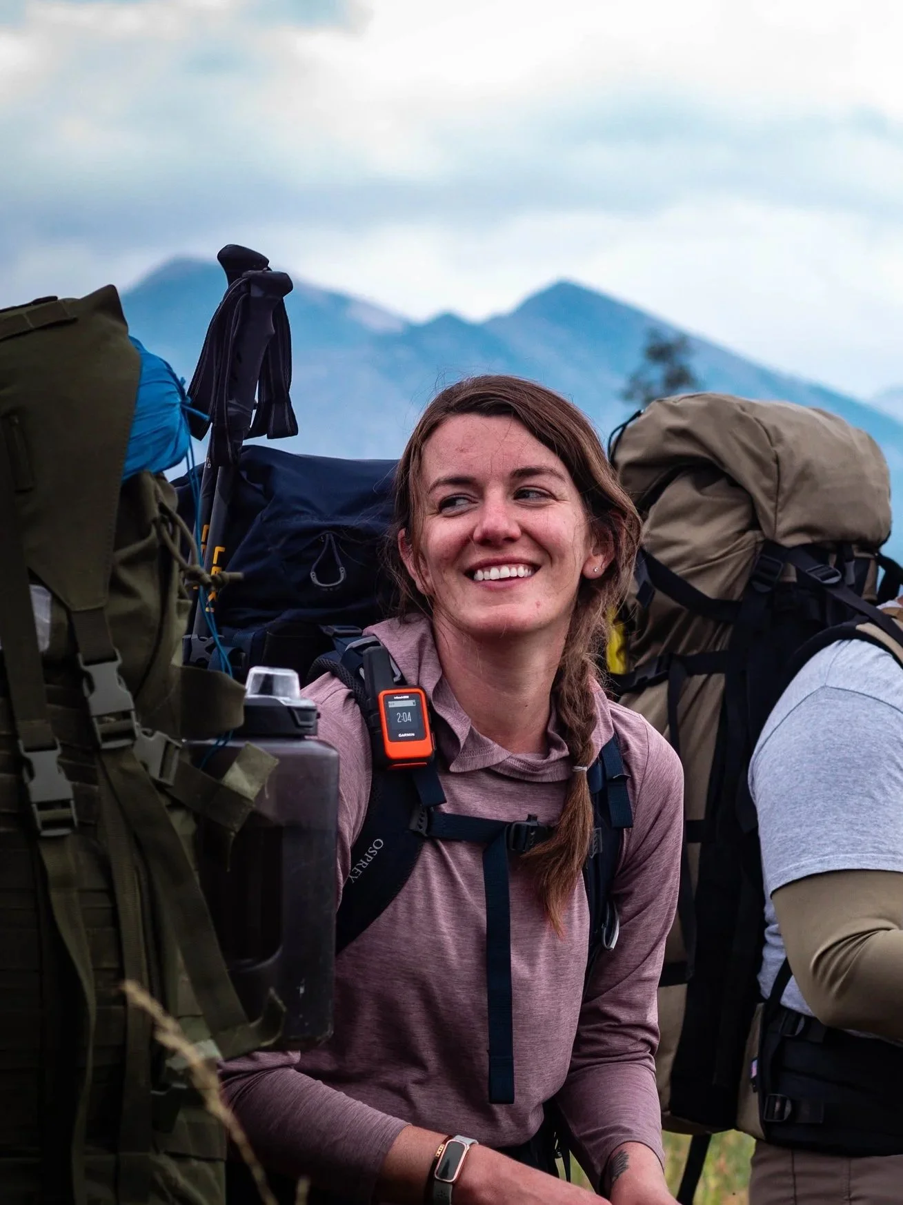 A woman smiles while hiking with a group in a mountainous outdoor setting, surrounded by backpacks and gear.