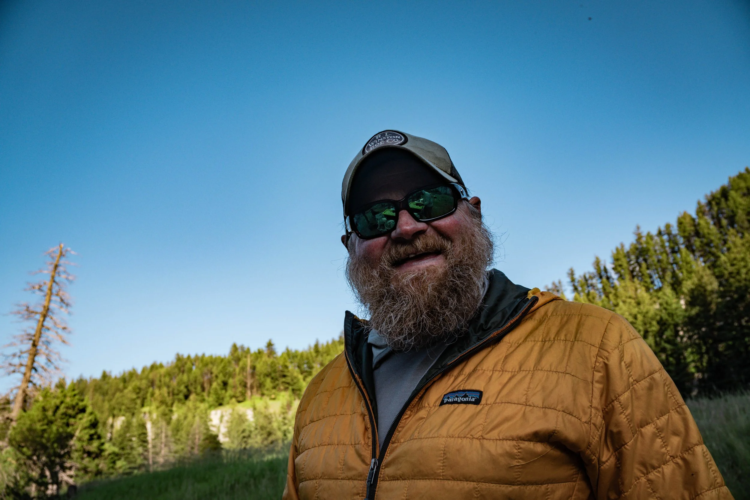 A man with a beard and sunglasses wearing a tan Patagonia jacket, outdoors in front of a landscape with trees and a lake under a clear blue sky.