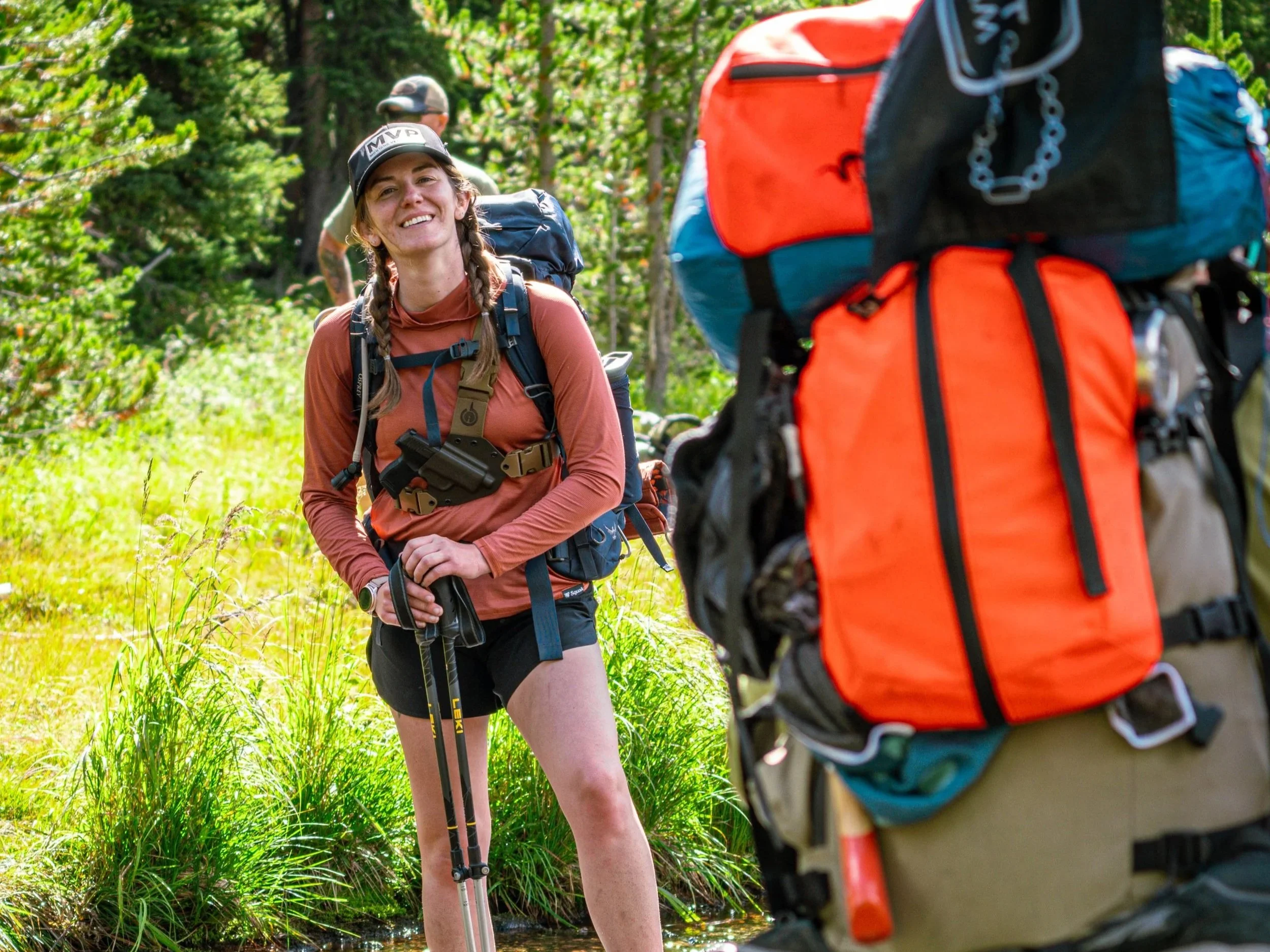 Smile of a woman hiker with braids and wearing a cap, standing in a lush green forest with backpack and hiking poles, smiling at a person with a large orange and blue backpack.