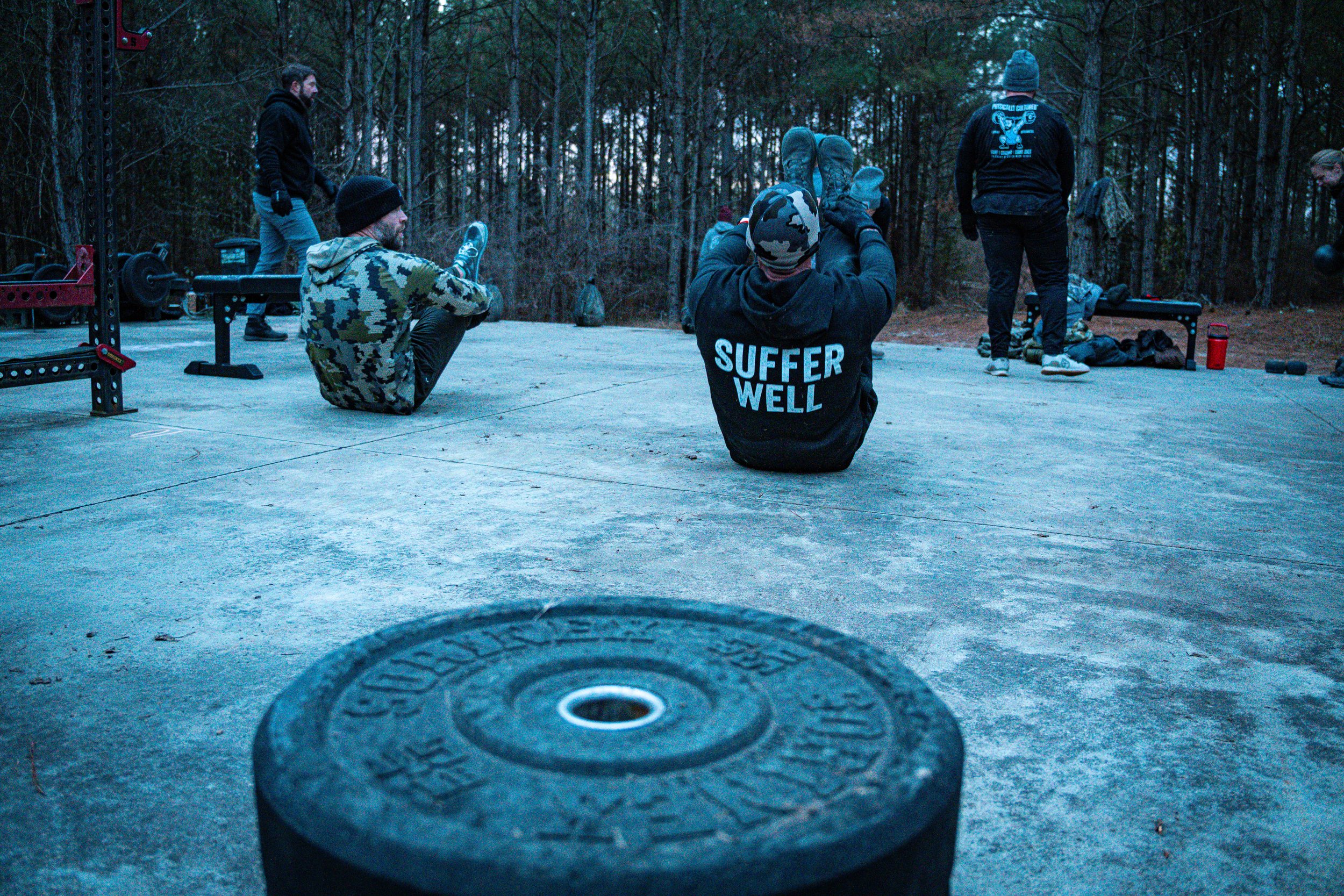 Group of people sitting and standing outdoors in a wooded area, some with gym equipment and weights, during dusk or dawn.