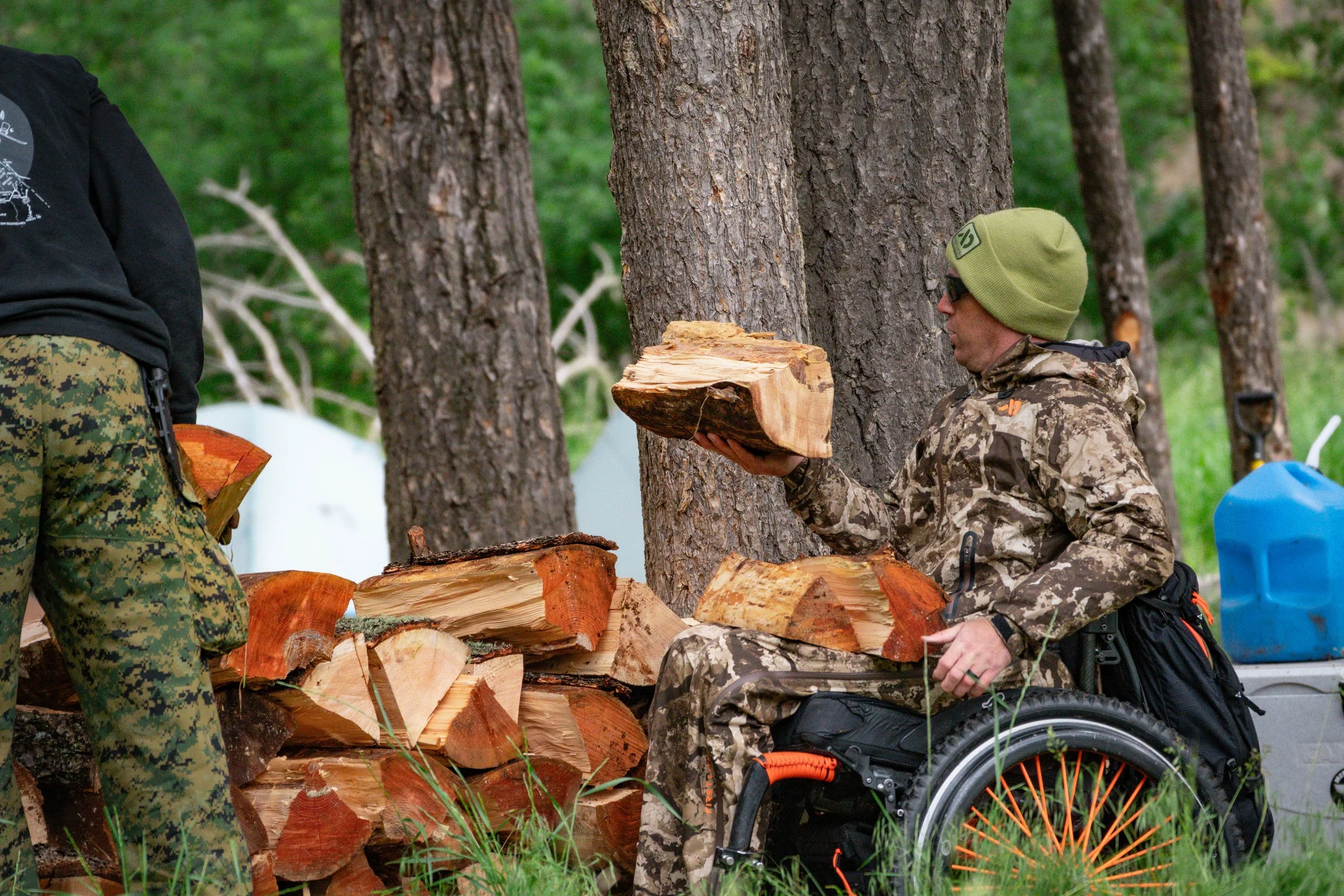 A person in a wheelchair wearing camouflage clothing and a green beanie, holding firewood, outdoors among trees with logs stacked nearby.
