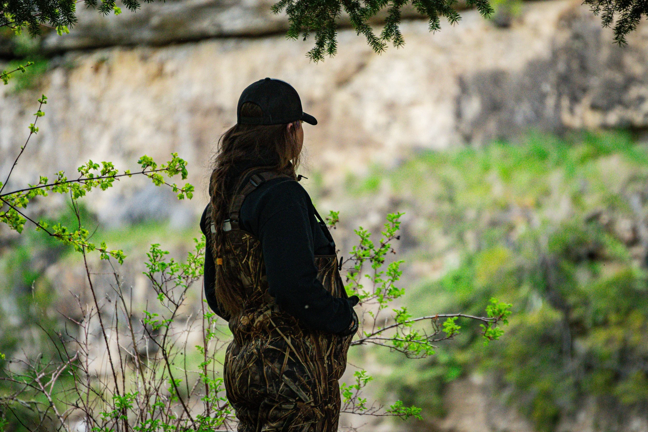 A woman with long hair wearing a black cap and camouflage clothing stands outdoors amidst green foliage, overlooking a rocky background.