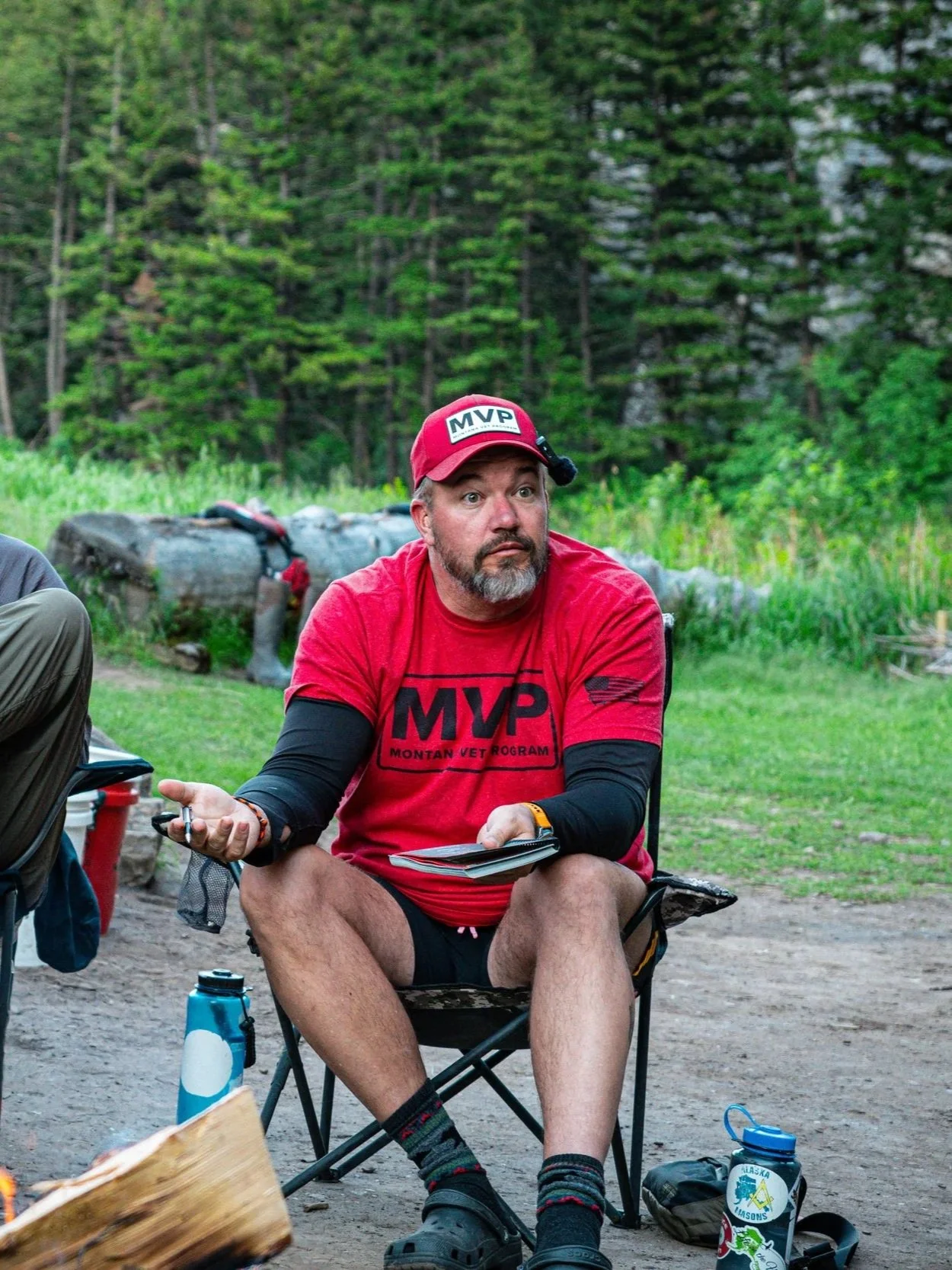 A man with a beard wearing a red MVP Montan Vet Program t-shirt, red cap, black shorts, and hiking boots, sitting outside in a forested area, holding a notebook, with a water bottle and camping gear nearby.