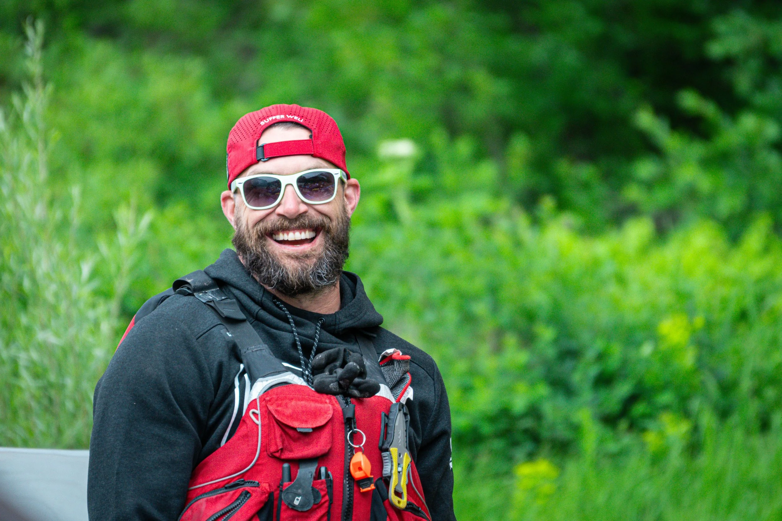 Smiling man with a beard wearing sunglasses, a red cap, and outdoor gear in a green natural setting.