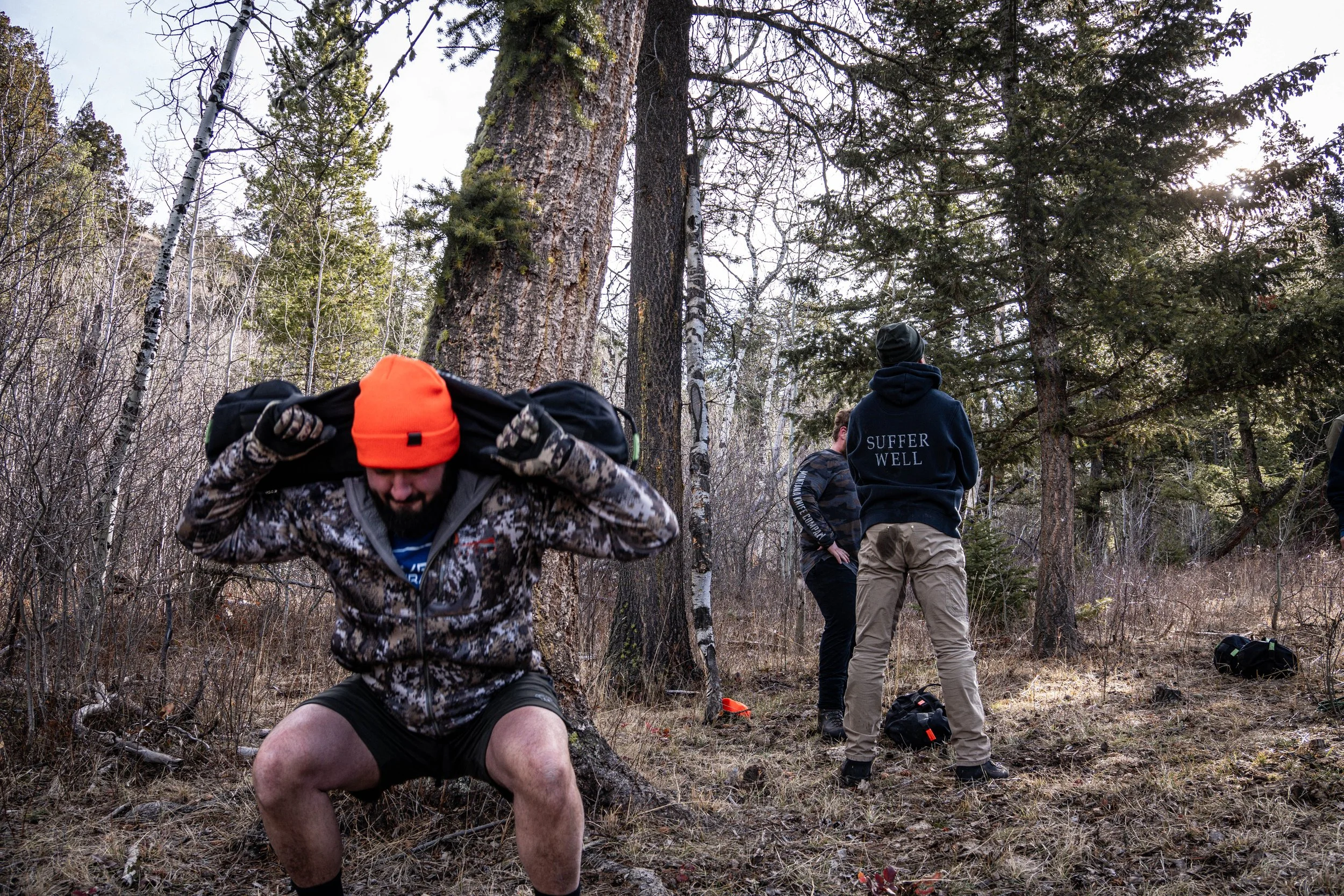 Three men in outdoor clothing and gear in a forest with tall trees and bare ground. One man in the foreground is crouching and adjusting his backpack. The other two men are standing, with one wearing a hoodie that says 'Suffer Well.' The sun is low, filtering through the trees.