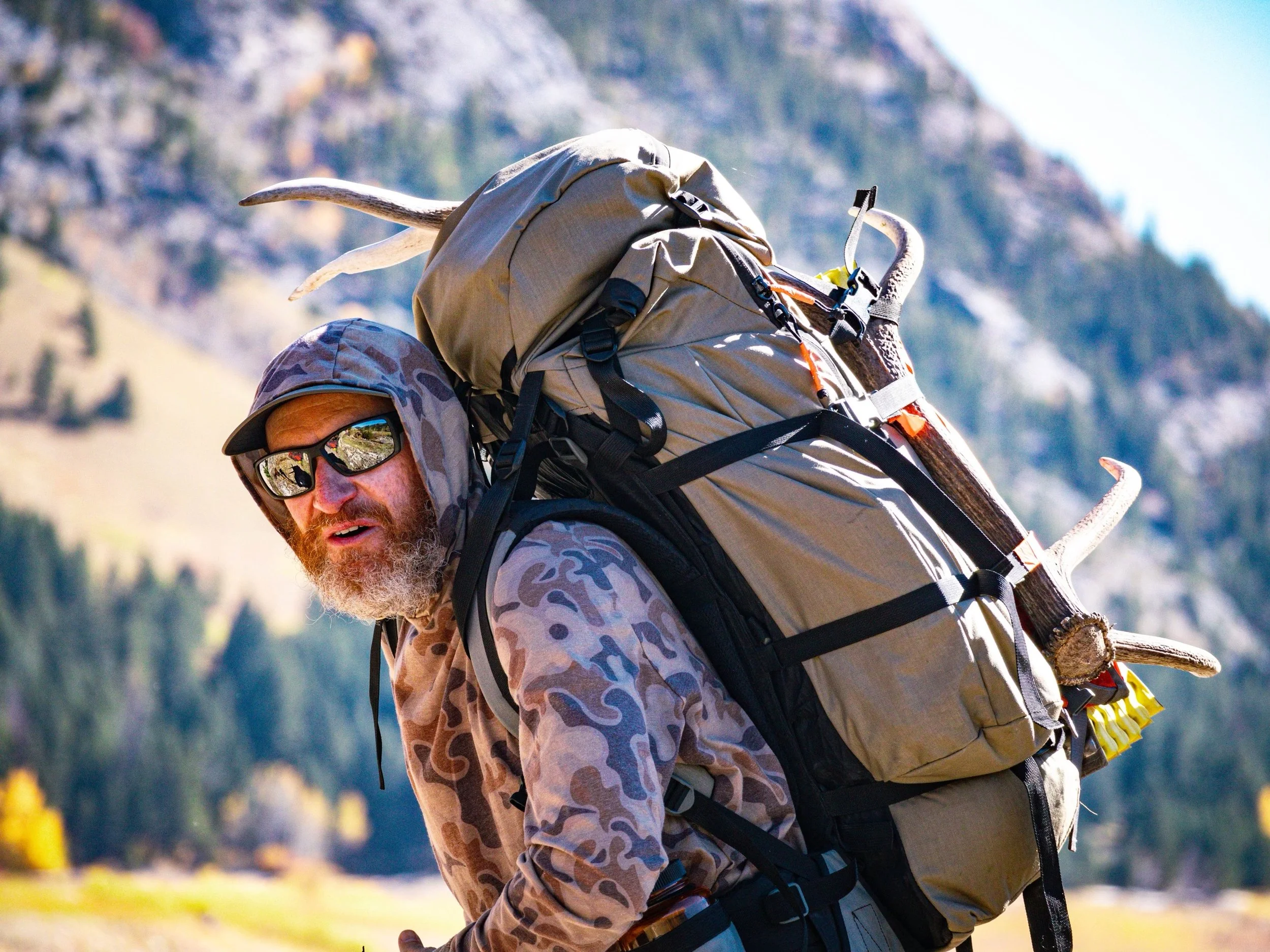 A man wearing a camouflage jacket, sunglasses, and a wide-brimmed hat with a camouflage pattern, carrying a large beige backpack with straps and gear, outdoors with a mountainous background.