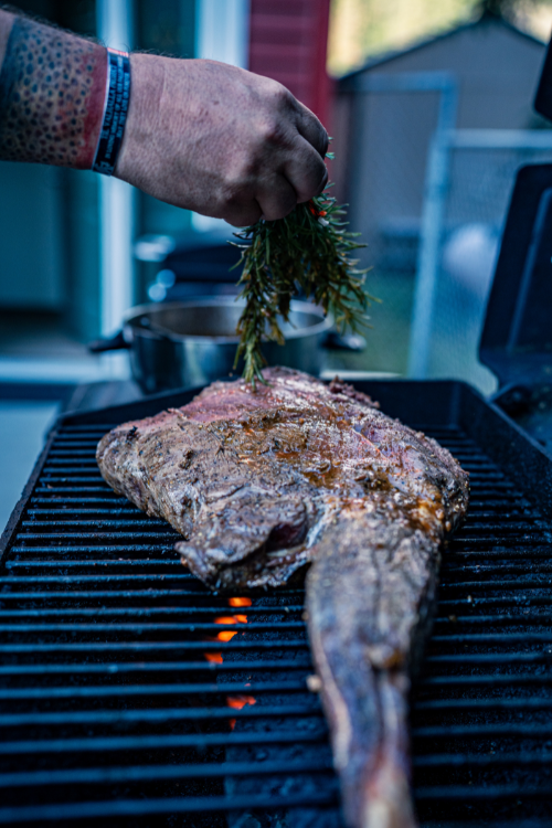 Person sprinkles herbs onto a large fish cooking on a grill outside.