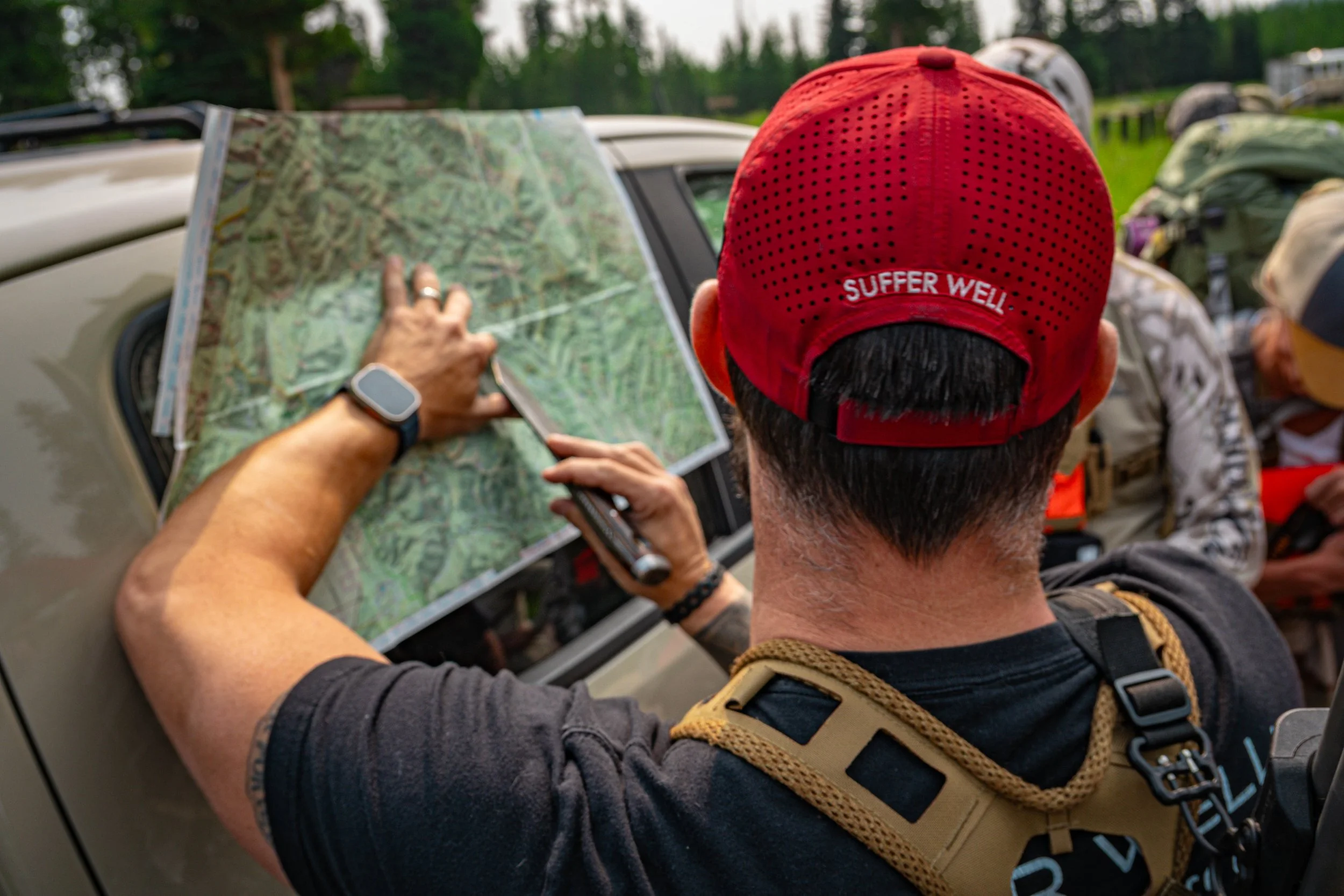 Back of a person wearing a red cap with the words "Suffer Well" looking at a map, with others in hiking gear nearby, outdoors in a wooded area.