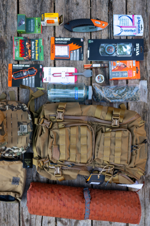 Outdoor gear and supplies laid out on a wooden surface, including a tan backpack, a rolled orange sleeping pad, water bottles, knives, flashlights, batteries, and emergency items.