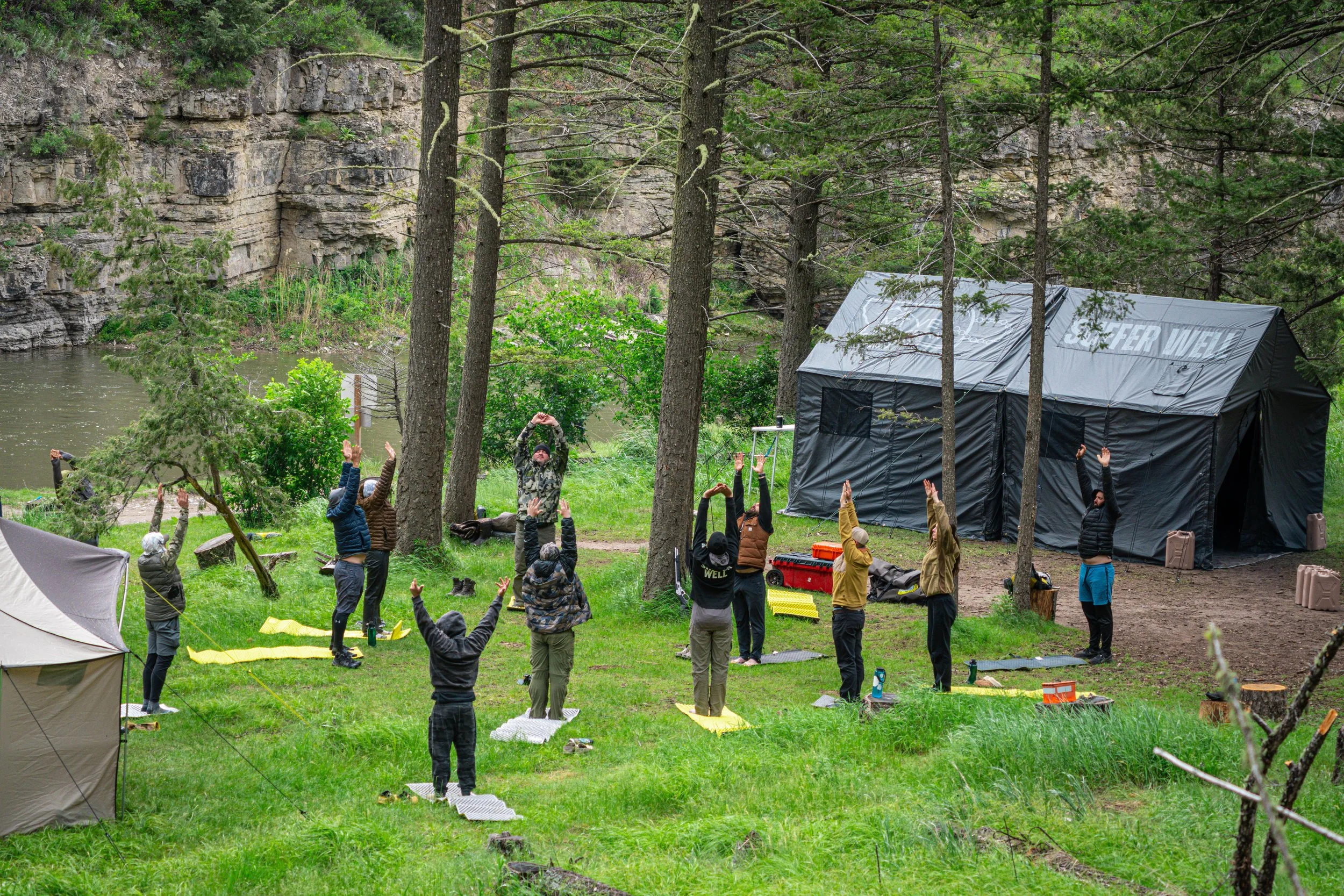People participating in outdoor yoga or stretching session near a river with trees and rocky cliffs in the background.